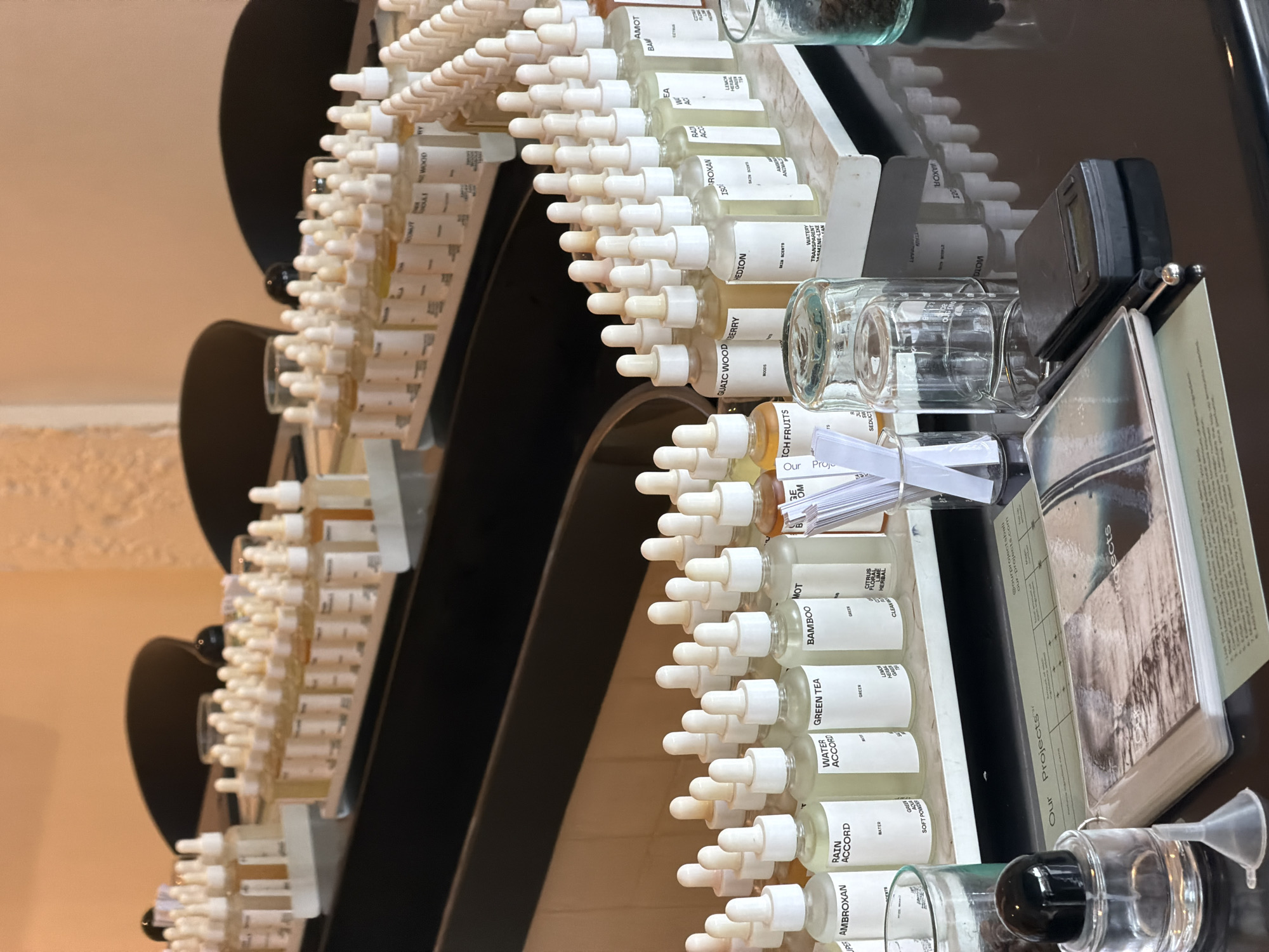 The image shows a close-up of a table with several rows of small glass bottles containing liquids, likely perfume or essential oils. These bottles are arranged on white trays. In the foreground, there are additional items on the black tabletop, including a notebook with the title "Our Projects," a small digital scale, a glass funnel, a round glass bottle with a black lid, and two clear glasses. The background is blurred, showing what appears to be a wall and chairs. The lighting is warm, suggesting an indoor setting. There are no people visible in the image. The scene appears to be in a laboratory or workshop setting, possibly for perfumery or scent creation. The mention of "Mengwi, Indonesia" in the prompt is not visually supported by any elements within the image itself.