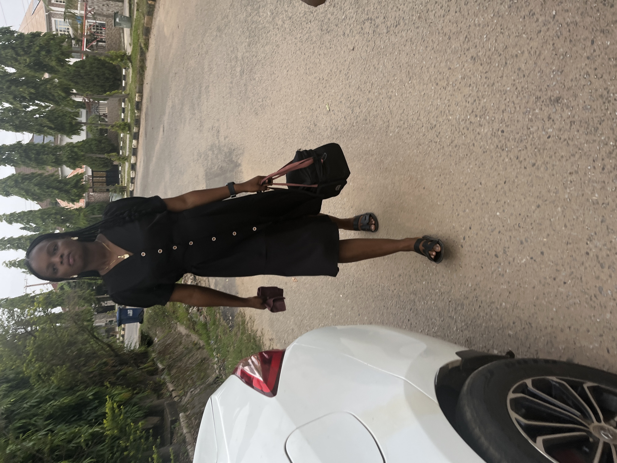 A young Black woman is captured walking or standing on a paved road under daylight conditions, likely in an urban or suburban area of Abuja, Nigeria. She is wearing a black v-neck dress adorned with white circular buttons down the front, dark sandals, and a thin gold chain with a small pendant. Her hair is styled in braids or dreadlocks. In her left hand, she carries a black bag with pink straps, and in her right, she holds a small reddish-brown object, possibly a cloth or wallet. A dark item, which could be a watch or bracelet, is visible on her left wrist. Her expression appears neutral as she looks towards the camera.
In the immediate foreground, the rear right section of a white car is partially visible, including its taillight and a portion of its wheel. The road surface is textured asphalt. To her left, a neatly maintained green verge with short grass is bordered by distinctive white and black striped curbs. Beyond the verge, a row of tall, slender evergreen trees lines the street, partially obscuring residential buildings in the background. Some structures with light-colored walls and dark roofs can be seen through and above the trees. A blue waste bin is also discernible amongst the background foliage. The lighting suggests a daytime setting, possibly with an overcast or hazy sky, as strong shadows are absent. No discernible text is visible in the image.
