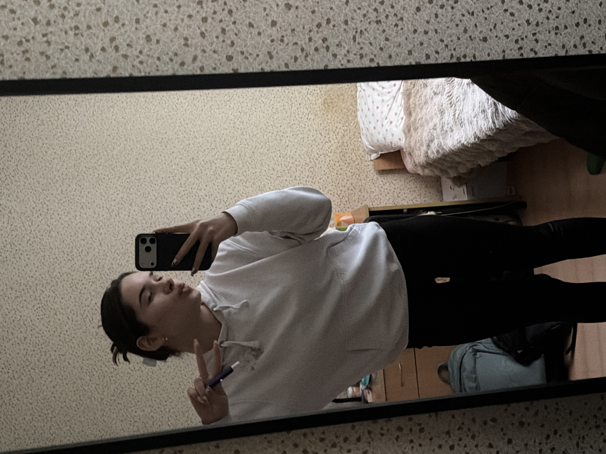 The image displays a young woman taking a mirror selfie indoors. She is reflected in a large mirror with a black frame, capturing her full body and parts of the surrounding room. The woman is wearing a white hooded sweatshirt and black pants. She holds a black smartphone in her left hand, using it to capture the reflection. Her right hand is making a "peace" sign (V-sign) and she is holding a small, dark purple cylindrical object, possibly a pen or vaping device. Her facial expression is a playful pout or kissy face.
The reflected background reveals a room with light-colored, textured walls characterized by small dark speckles. To the right of the woman, a bed is visible, covered with a soft, white, fluffy blanket and a pillow with a white base and red/pink polka dots. Beneath the bed, a light blue backpack or bag rests on a wooden floor. A small green object, possibly a slipper, is partially visible on the far right. The lighting in the room appears to be ambient, without strong natural light, creating a casual and relaxed atmosphere. No discernible text is visible within the image. The location is understood to be in Sofia, Bulgaria.