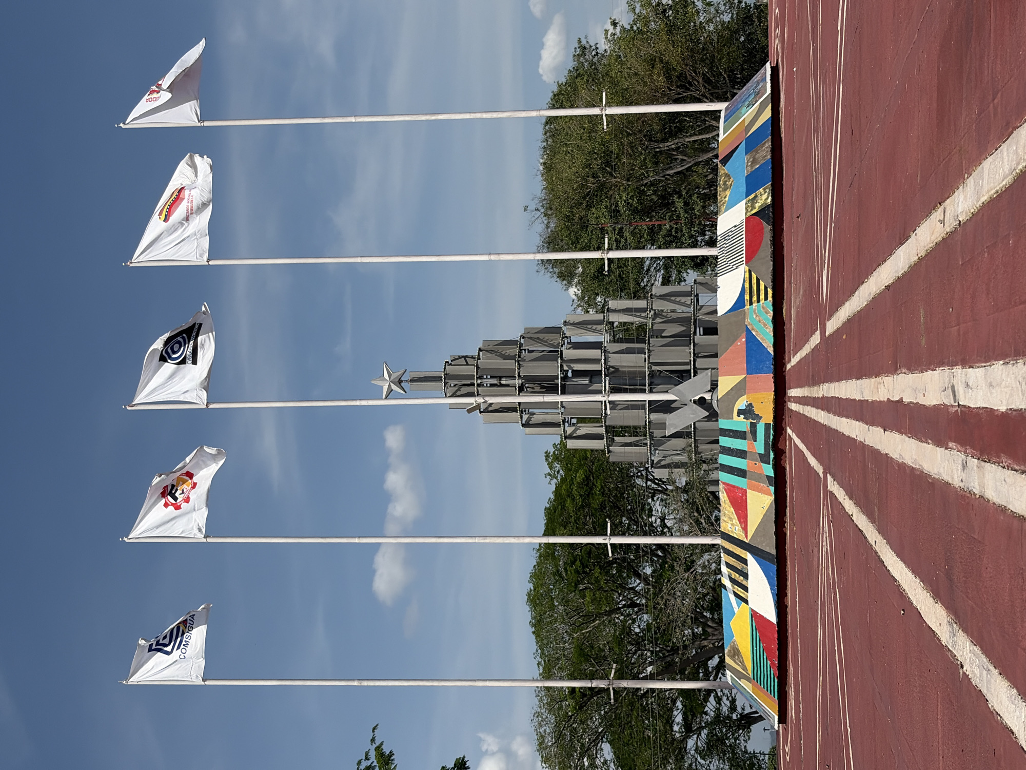 An outdoor scene in Ciudad Guayana, Venezuela, captured on a bright, sunny day under a clear blue sky with scattered white clouds. The image is oriented vertically (originally landscape, rotated clockwise), depicting a vibrant public space with various elements.
In the foreground (bottom when correctly oriented), a reddish-brown ground surface, possibly a track or a painted wall, stretches across the bottom, adorned with converging white lines that create a sense of depth. To the far left of this surface, a multi-colored wall section displays an abstract geometric pattern with blocks of red, blue, yellow, black, and white.
Dominating the mid-ground are five white flags, each flying from a tall white flagpole, arrayed horizontally across the frame. From left to right, the flags bear distinct logos and text:
1. A red logo with the word "BODIM".
2. A red hot dog graphic with text that includes "VENEZUELA".
3. A dark, circular, eye-like logo, with partially obscured text that appears to be "REVOLUTION SPORTS".
4. A red and yellow emblem, possibly featuring a figure within a cog or gear.
5. A blue geometric logo with the word "COMSIGUA".
Behind the flags stands a prominent metallic monument, resembling a modern, abstract Christmas tree or pyramid, constructed from stacked silver-gray panels and topped with a five-pointed star. Lush green trees are visible in the background, providing a natural contrast to the man-made structures. The bright sunlight indicates it is daytime, likely morning or early afternoon. The scene conveys a public or corporate display, possibly within a park or event area, highlighted by the various company or organizational flags.