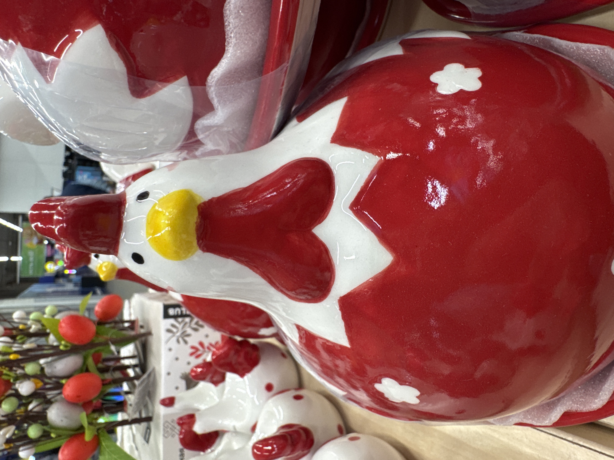 The image shows a collection of red and white ceramic chicken-shaped containers, possibly for food storage or decorative purposes. These are displayed on a wooden shelf, likely in a retail setting. Visible to the left are some decorative branches with red and white spherical items, possibly berries or eggs, suggesting an Easter or spring theme. The containers have a glossy finish and feature white accents like flower-shaped designs. Some are covered in clear plastic or cellophane. The background indicates a store environment, and considering the prevalence of Easter decorations, the scene could be taking place in a department store, craft store, or grocery store during the spring season. The image is not enough to specifically place it in Saint Petersburg, Russia.