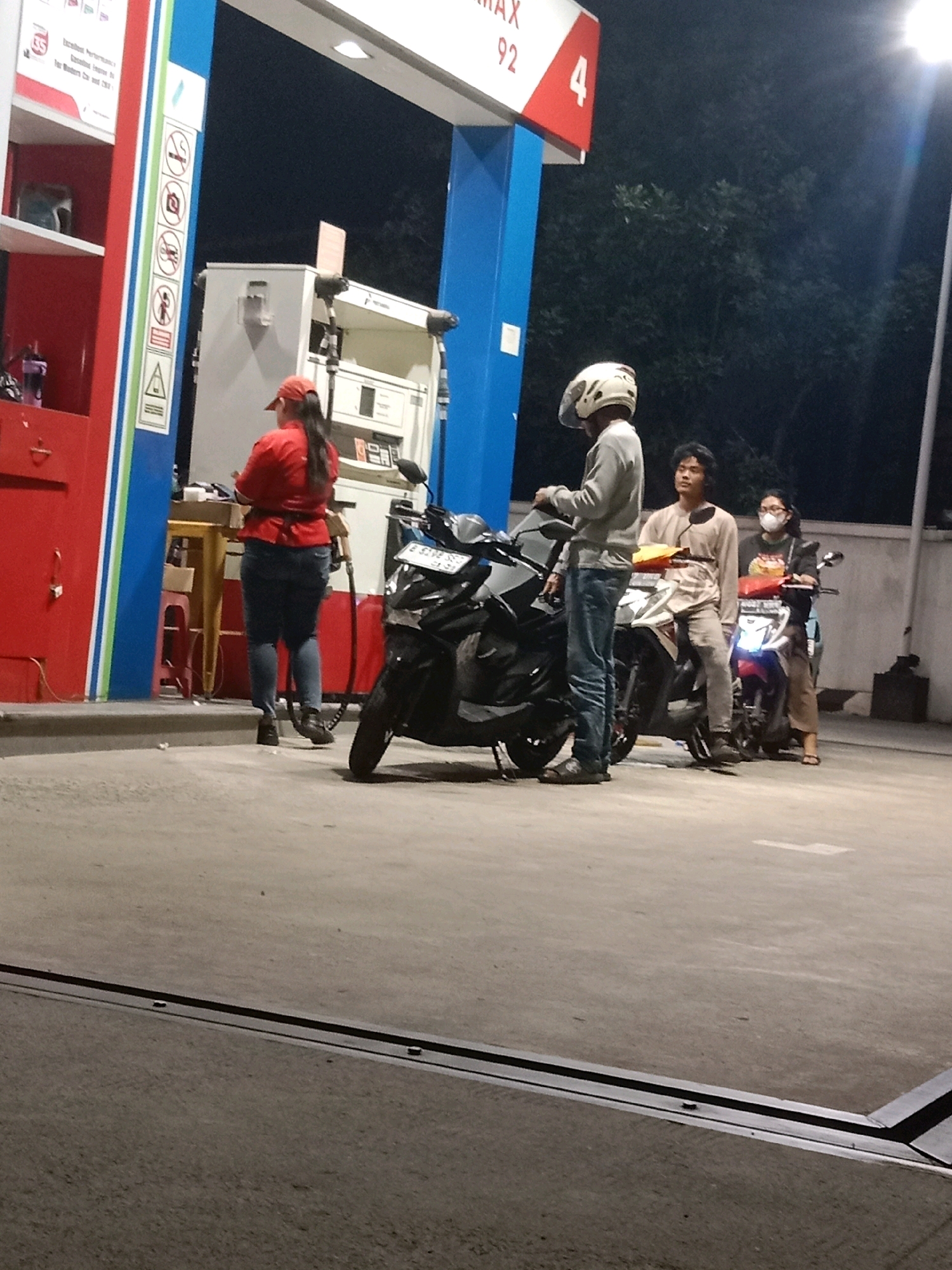 The image depicts a scene at a gas station in South Tangerang, Indonesia, during nighttime. A female gas station attendant wearing a red uniform and cap is attending to a pump. Three individuals on motorcycles are present. One individual, wearing a helmet and a light-colored long-sleeved shirt and jeans, stands near a black motorcycle. Another individual, a male with dark hair and wearing a light brown patterned long-sleeved shirt and pants, sits on a white motorcycle. A third individual, a female wearing glasses, a mask, and a dark-colored long-sleeved shirt with a graphic design, sits on a motorcycle with a red front. The gas station features a blue and white structure with a sign displaying "MAX 92" and the number "4". The ground is paved concrete. Trees are visible in the background.