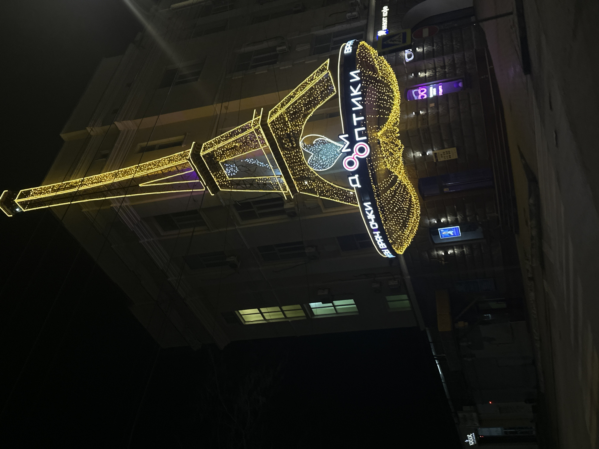 A nighttime street scene in Rostov-on-Don, Russia, features a large illuminated sign. The sign is constructed from yellow and white lights, depicting a pair of eyeglasses and the Russian text "Дом Оптики" (House of Optics). The sign is positioned in front of a multi-story building. Other signs are visible on the adjacent building. The street is dark, and trees are visible at the bottom of the frame.