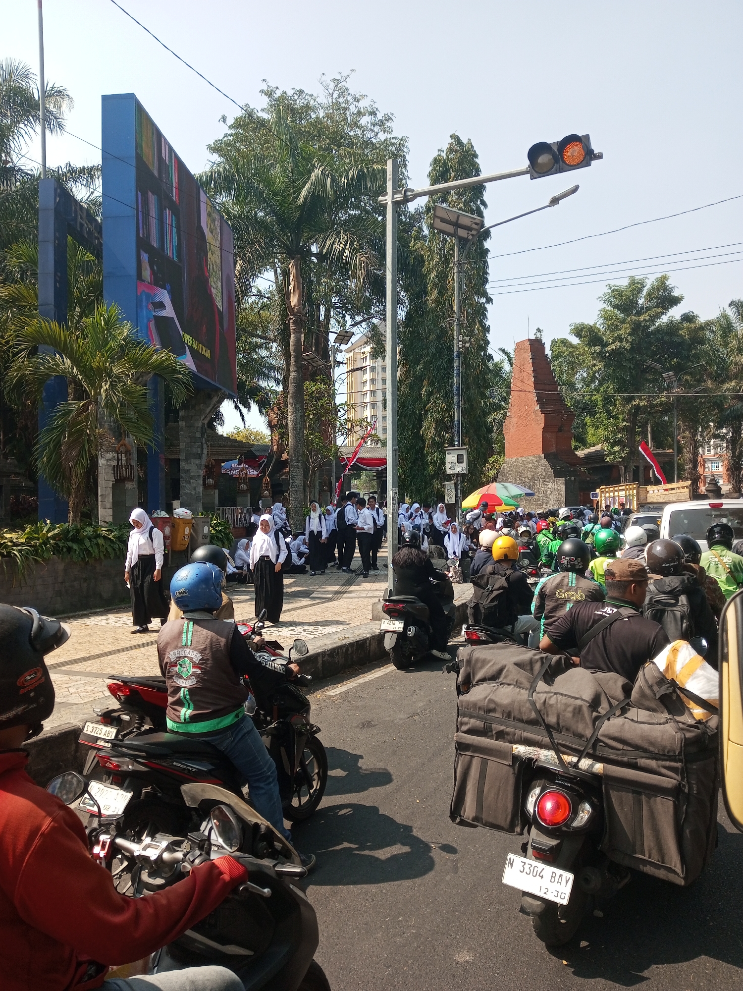 The image captures a street scene in Malang, Indonesia, with a focus on a group of students and a traffic jam. In the foreground, two men on motorcycles are stopped, one wearing a red jacket and the other a black jacket with a green trim. Behind them, a line of motorcycles forms a traffic jam, with many riders wearing green or black vests, suggesting they might be delivery personnel.
On the sidewalk to the left, several students in white shirts and black hijabs or pants are gathered. Some are standing, while others are sitting. They appear to be waiting or observing the traffic. Further back, more students in similar attire are visible, along with some in white shirts and black pants, and others in white shirts and black skirts.
A large digital billboard displaying what looks like book covers is prominent on the left side of the image. Behind the students and the billboard, palm trees and other lush vegetation can be seen. A tall, narrow brick structure, possibly an ancient monument or gateway, stands further down the street, with trees surrounding it. A traffic light with an amber light illuminated is visible against the clear sky. The overall atmosphere suggests a busy urban environment, possibly near a school or educational institution, with the ongoing traffic presenting a common urban challenge.