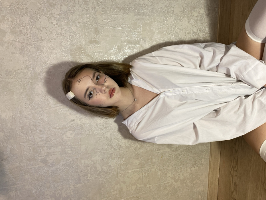A young woman lies sideways on a textured surface, partly on a wooden floor. She wears a large white shirt and white socks. Her hair is shoulder-length and brown. She has makeup on her face that looks like cracks on a doll. A small white flower clip is in her hair. She wears a delicate necklace with a small diamond-like pendant. Her expression is neutral, and she gazes directly at the viewer.
