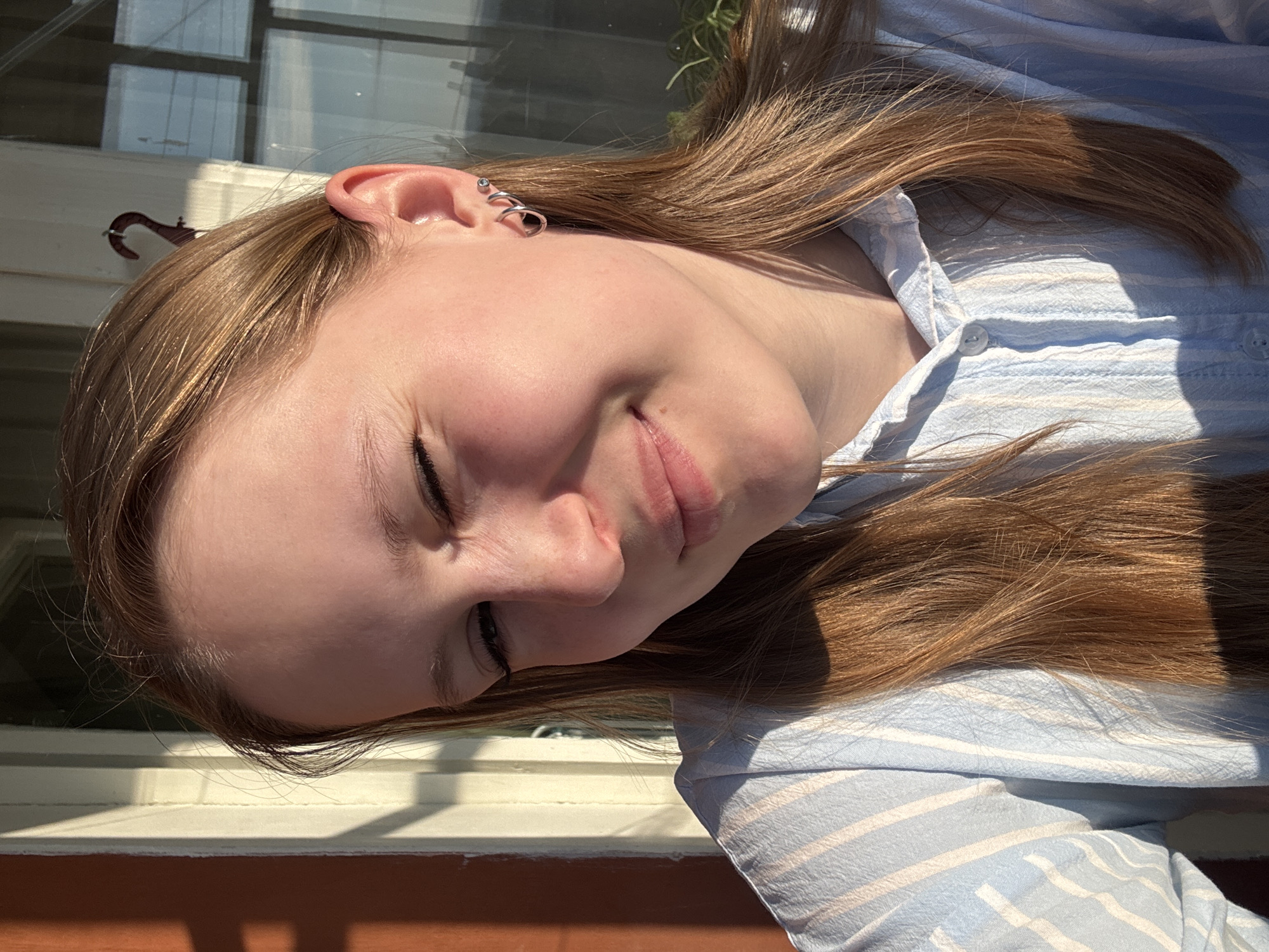 A young person with light brown hair is facing downward towards the lower left corner of the image. They are wearing a light blue, striped shirt. Multiple helix piercings are visible on their left ear. The background includes a window frame and what appears to be an exterior wall. There is not enough information to place this image in the city of Chaikovsky, Russia.