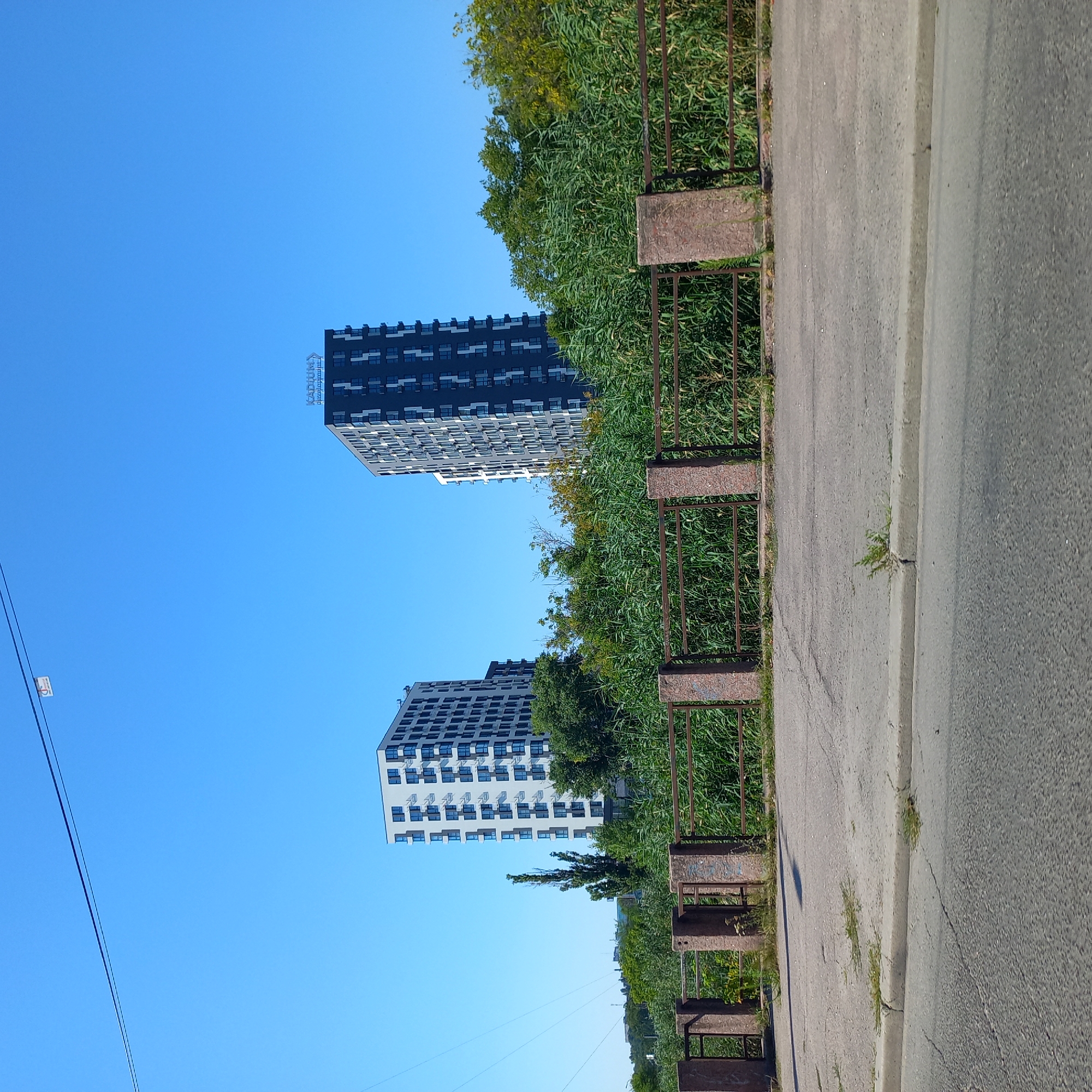 This image depicts two modern high-rise buildings against a clear blue sky. The building on the left is predominantly white with dark blue window frames and balconies. The building on the right is darker, with a gray facade and white window frames. A sign with blue lettering reading "KADIUM" is visible at the top of the right building, suggesting it may be a residential complex or development. In the foreground, there is a metal fence with stone pillars, backed by dense green vegetation. A paved road and sidewalk are visible in front of the fence. The scene appears to be located in an urban environment, possibly near a waterfront given the lush vegetation. Based on the "KADIUM" sign, this is likely the Kadium residential complex in Kropyvnytskyi, Ukraine, situated near the Inhulets River. There are no people or vehicles visible in the image.