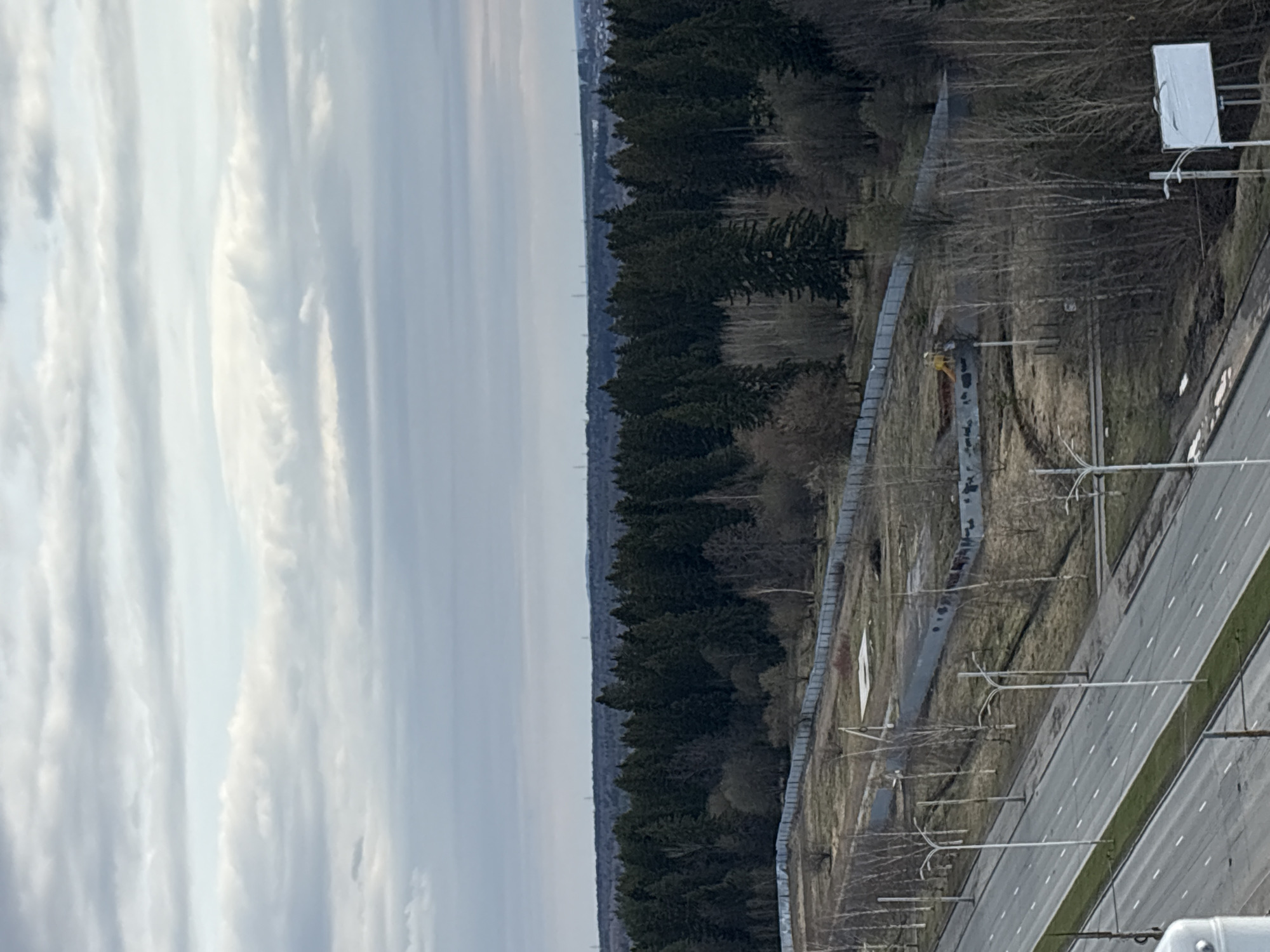 The image shows a view overlooking the city of Izhevsk, Russia. The sky is cloudy with some patches of blue visible. A dense forest line runs through the middle ground of the image. Adjacent to the forest is a cleared area with a gray fence and sparse vegetation. A multi-lane highway with overhead power lines runs parallel to the cleared area and forest.