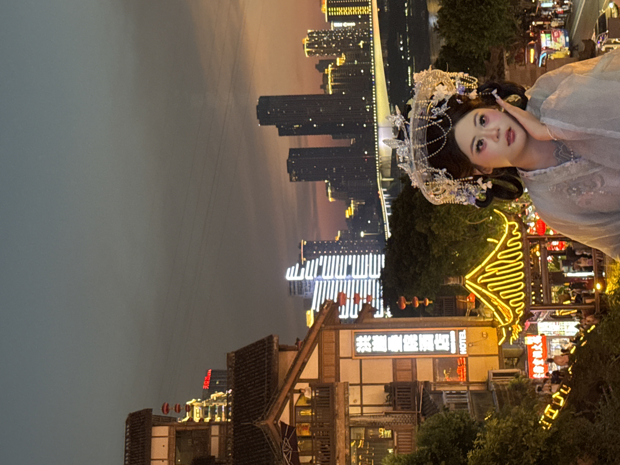 An outdoor medium shot captures a woman in traditional Chinese attire and elaborate headwear posing in front of a cityscape at dusk. The woman is positioned in the lower right quadrant of the frame, with her face turned to the left and her right hand resting on her cheek. She has dramatic makeup with pink eyeshadow and blush. Behind her, the cityscape unfolds with traditional Chinese architecture in the foreground on the left, featuring a multi-story building with a tiled roof and illuminated windows, and a sign that reads "HOTEL." Further back, modern skyscrapers rise against the twilight sky, with one particularly striking building illuminated with horizontal white lights. A bridge spans a body of water in the mid-ground, also adorned with lights. The scene suggests a location in Yuzhong District, China, known for its blend of historic and contemporary architecture, and its vibrant nightlife. The atmosphere is festive, with decorative lights adorning the traditional buildings and surrounding foliage.