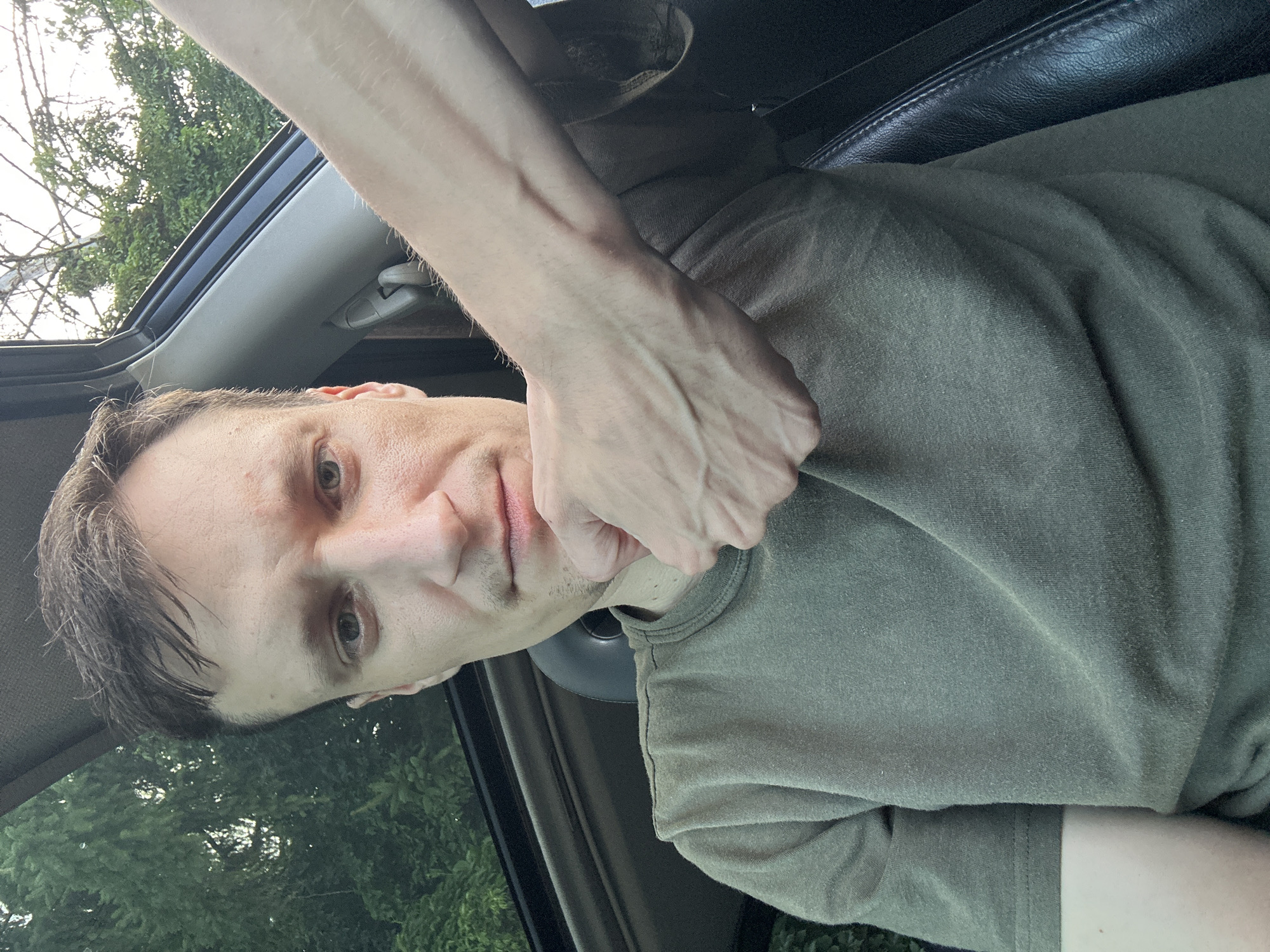 The image shows a close-up, selfie-style shot of a man in a car. He is wearing a green t-shirt and his right arm is extended, with his hand resting on his chin. His left arm is also visible, resting on his shoulder. The man has short brown hair and is looking directly at the camera. The interior of the car is visible, including a dark-colored seat and part of the sunroof. Outside the car, green trees and foliage can be seen, suggesting an outdoor, possibly rural or suburban, location. The lighting suggests it is daytime. There are no visible indicators that this photo was taken in Shatava, Ukraine.