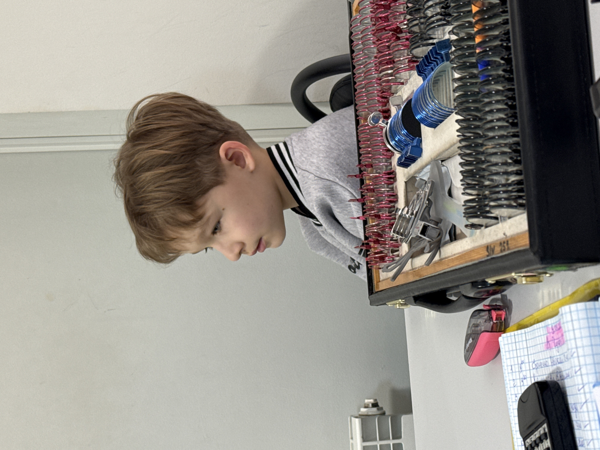 A boy with light brown hair is looking down at a container holding various metal springs. The container is resting on top of a white surface on which there is also a notebook, a pink pencil sharpener, and a black calculator. The background consists of a white wall. The location is unknown.