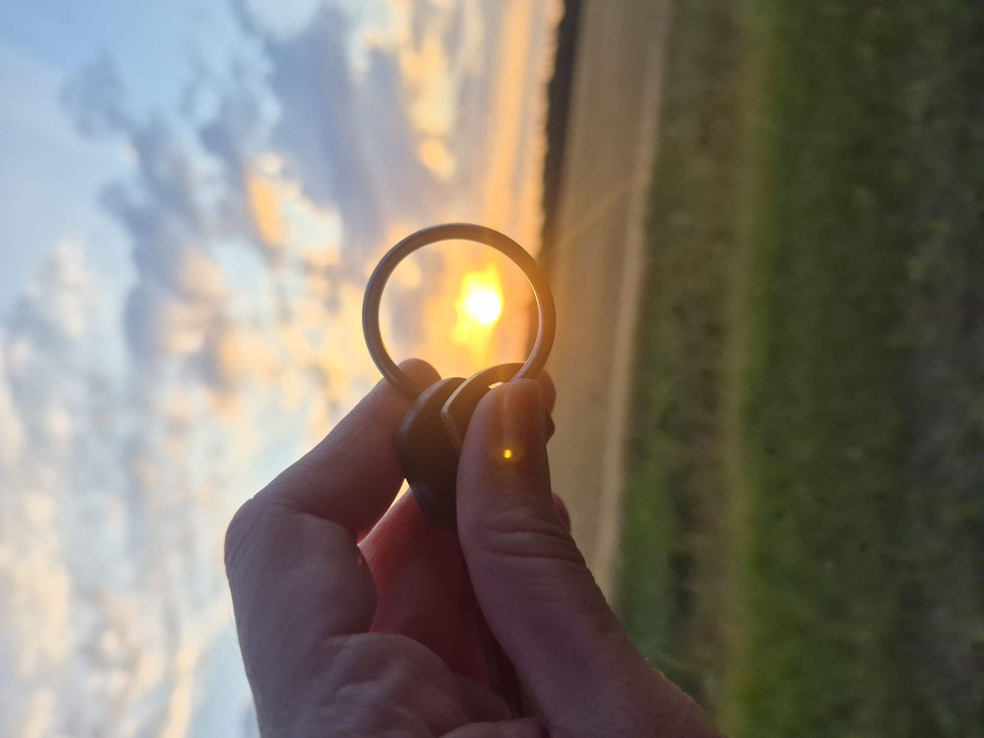 A close-up, low-angle shot shows a hand holding a keychain with two dark, smooth objects and a silver ring against a sunset sky. The sun is visible through the ring, casting a warm glow on the scene. The background features a blurred landscape of green fields and a distant treeline under a sky filled with clouds painted in hues of orange, yellow, and blue. The image was captured in Sidorovy Gory, Russia.
