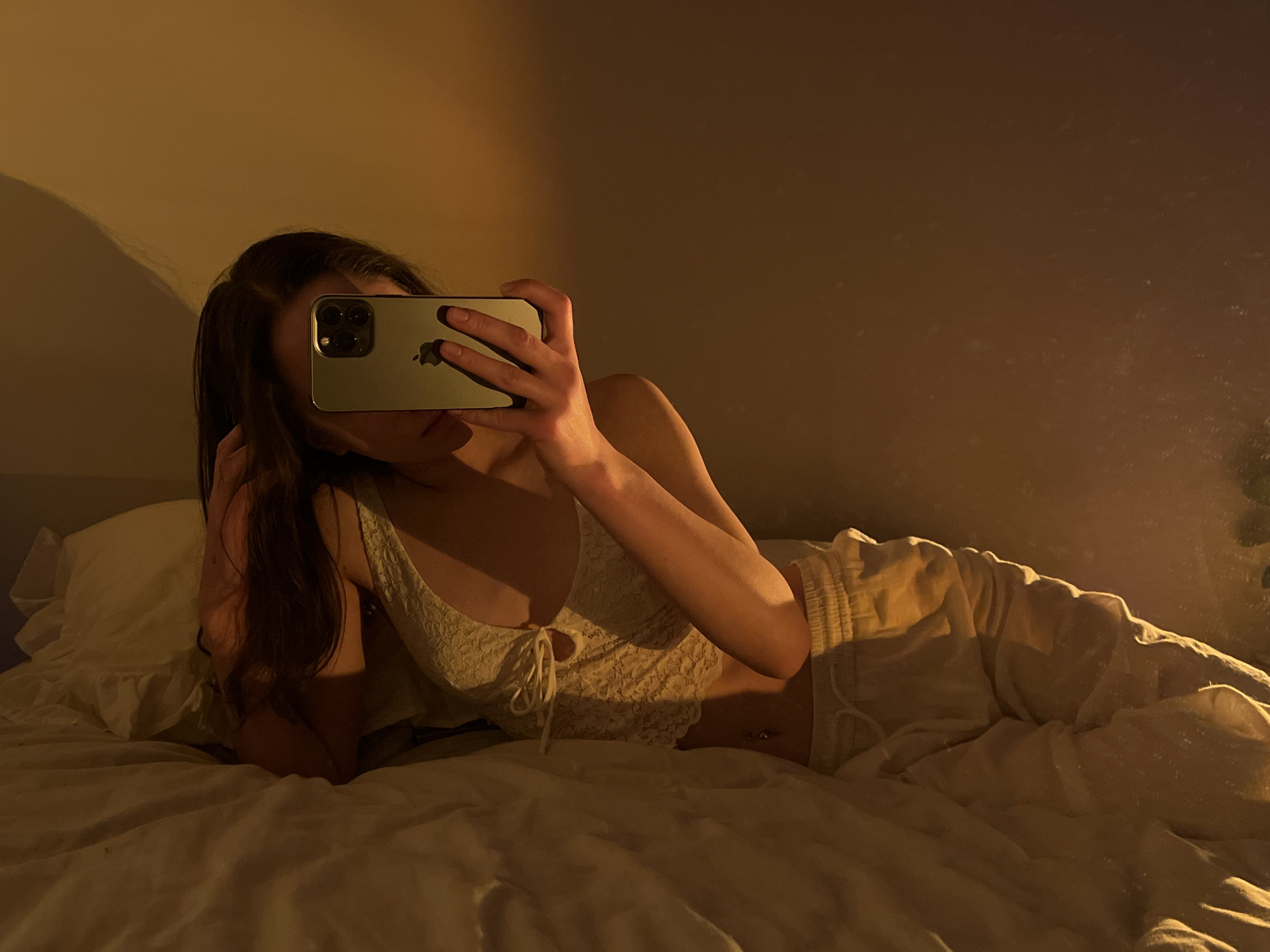 A person is lying on a bed, taking a selfie with a phone. The person is partially obscured by their phone and hair, but appears to be wearing a light-colored, lace top and light-colored pants. The bed is covered with white linens, and there are pillows. The room is dimly lit, with a warm, yellow light casting shadows on the wall. The image does not contain any information that would allow for the determination of the location in Ternopil, Ukraine.