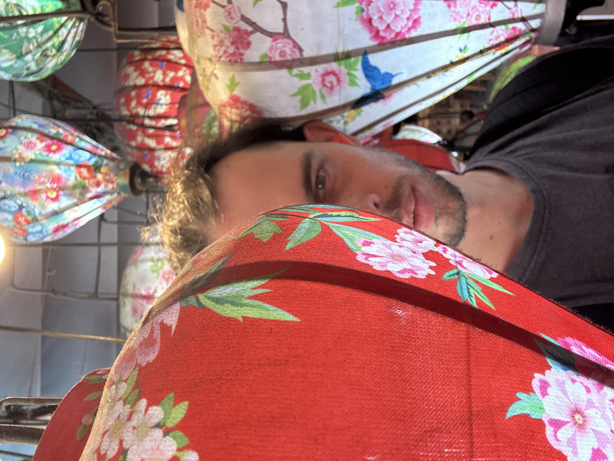 The media file is an eye-level, close-up, portrait-oriented shot of a man lying down, with his head and upper body dominating the frame. He is looking directly at the camera, with a neutral expression. The background is filled with traditional Vietnamese lanterns of various colors and floral patterns. Some lanterns are green, red, and white, adorned with pink flowers and blue birds. The man is wearing a dark gray t-shirt. The foreground is occupied by a large, red lantern with white and pink floral patterns, draped across the lower part of the image. The scene appears to be indoors or in a covered area, possibly a market or a shop selling lanterns. The lighting is soft, suggesting it could be daytime or the lanterns themselves are providing a gentle illumination. The overall impression is intimate and personal, with the man's gaze creating a direct connection with the viewer. The presence of numerous decorative lanterns strongly suggests the setting is in Đà Nẵng, Vietnam, known for its vibrant lantern culture, especially during festivals.