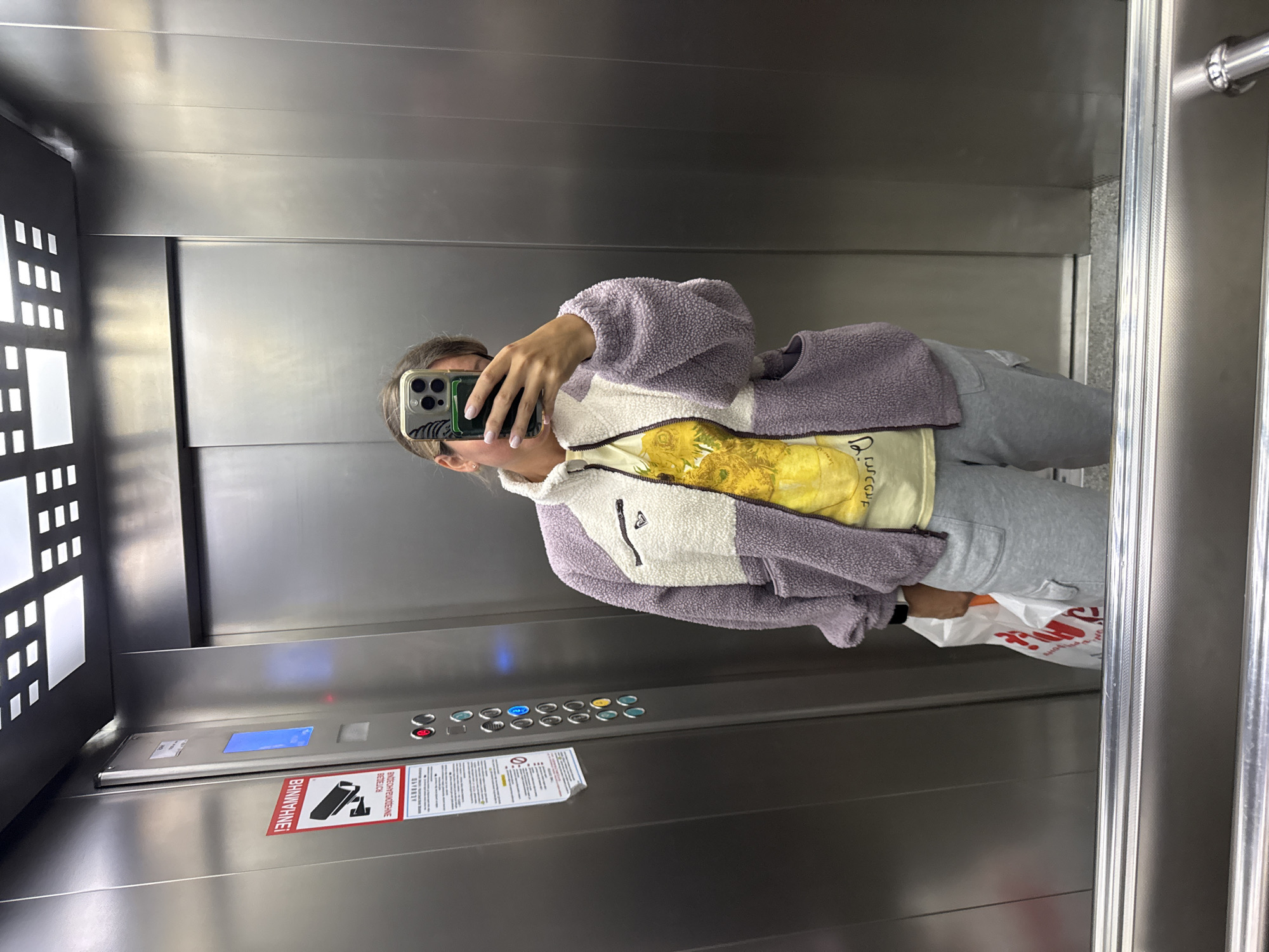 A person is taking a mirror selfie inside an elevator. The elevator has brushed stainless steel walls and control panel. The control panel is labeled in Russian. The elevator is equipped with a digital floor display and has illuminated wall decor. The location, Sochi, Russia, is inferred from the presence of Russian language signage. The person is wearing gray sweatpants, a yellow t-shirt, and a white and purple fleece jacket. They are holding a shopping bag with red and white text. The phone being used to take the picture has a green card holder attached.