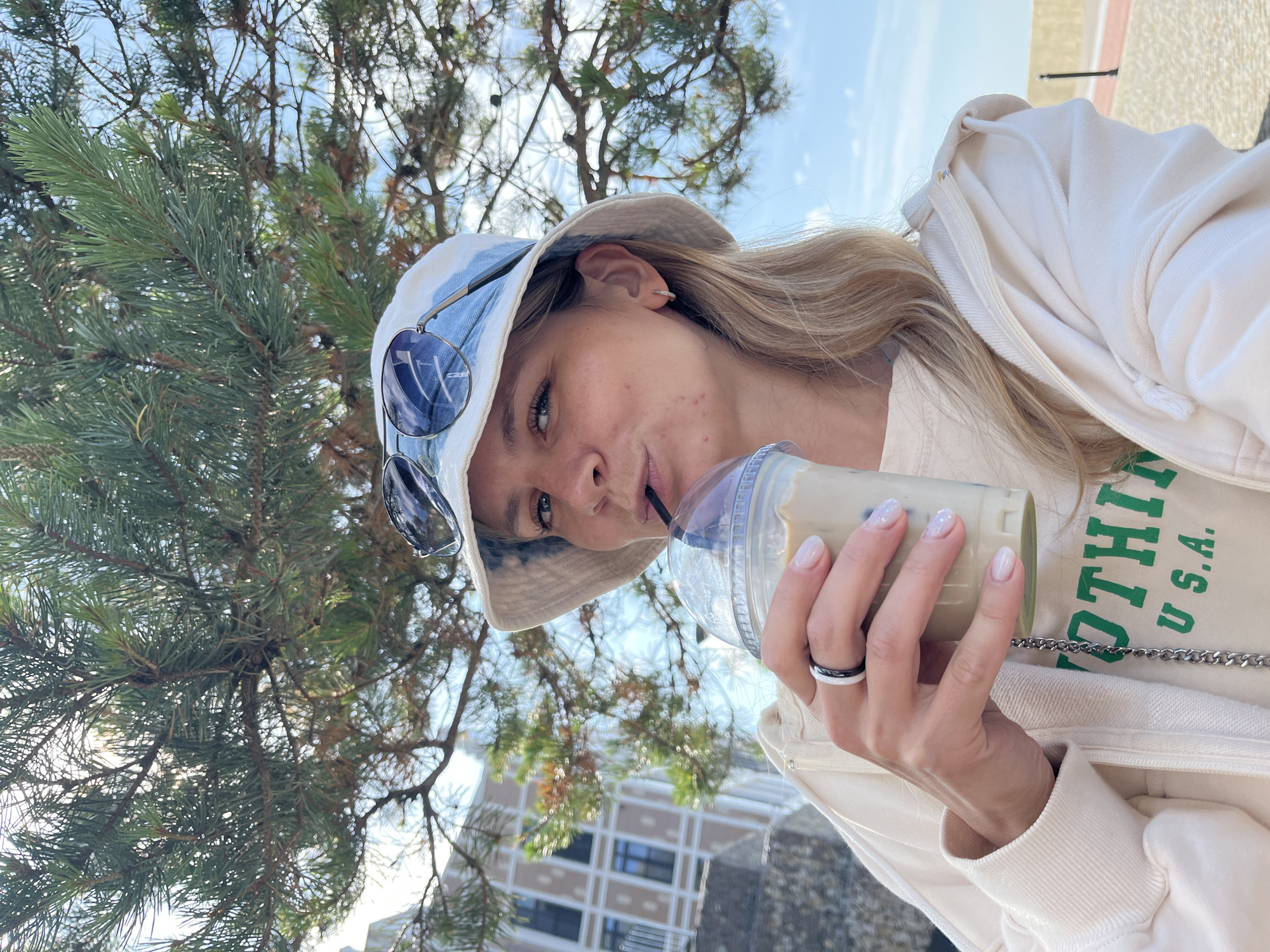 An outdoor, medium shot, selfie-style photograph captures a young woman with blonde hair and fair skin enjoying a beverage on a sunny day. She is wearing a light-colored bucket hat with a tie-dye pattern and blue tinted sunglasses pushed up on her head. A light-colored hoodie is visible, with a cream-colored t-shirt underneath featuring green lettering that reads "NOTHING U.S.A.". The woman is holding a clear plastic cup with a straw, from which she is drinking a light-colored beverage, likely iced coffee. She has a ring with white and black bands on her left hand and has light-colored nail polish on. Her right hand is holding the cup.
In the background, there is a large pine tree with green needles and branches obscuring the sky in the upper portion of the image. To the left, a multi-story building with a light-colored facade and dark windows is visible. The lower part of the background shows a sandy area with a paved pathway and a black post. The overall setting suggests a park or a promenade area in Svetlogorsk, Russia, possibly near the Baltic Sea, given the typical coastal vegetation and architecture found in the city. The lighting indicates it is daytime.