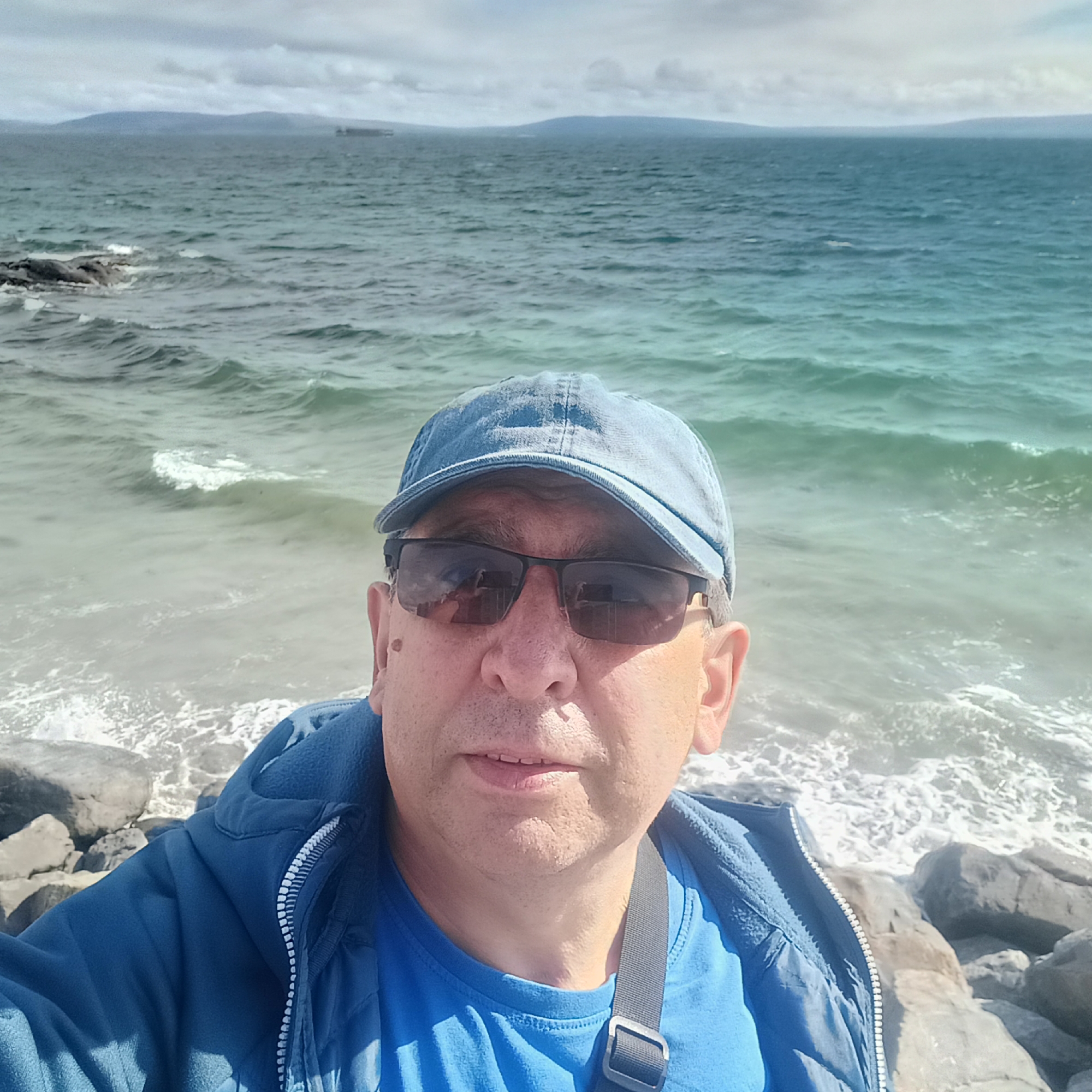A man wearing a blue baseball cap and sunglasses takes a selfie on the rocky shore of Galway Bay in Galway, Ireland. The ocean, with turquoise and grey waves, stretches out behind him, meeting a cloudy sky. In the distance, a hilly coastline is visible. The man is wearing a blue jacket over a blue t-shirt. The waves are crashing against the rocks at the edge of the water.