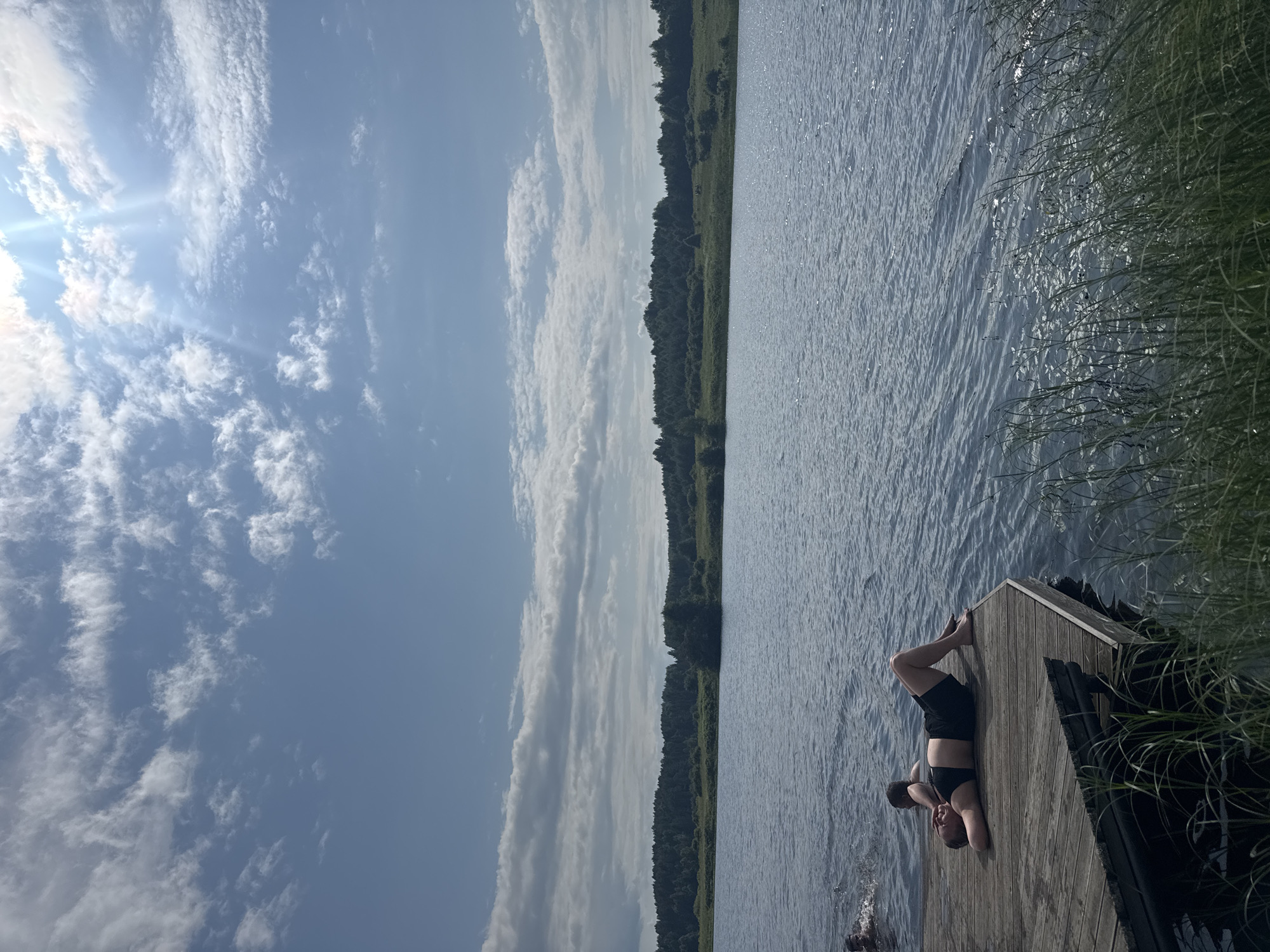 The media file shows a person lying on a wooden dock by a body of water. The person is wearing black shorts and appears to be relaxing or sunbathing. The water is calm with small ripples, and the sky is blue with scattered white clouds. A line of trees is visible along the distant shore. The overall scene suggests a peaceful outdoor setting, possibly during the daytime given the bright sky. The media file is oriented vertically, with the sky occupying the left side and the water and dock on the right.