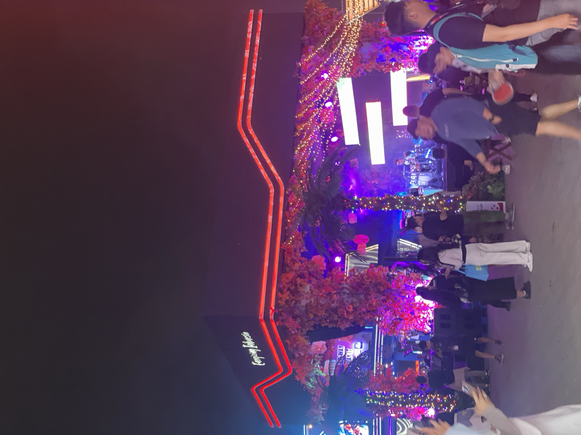 The image captures an outdoor event at night in Salembaran, Indonesia. A building with red neon outlining its architecture is visible on the left, bearing the name "Capjaen Indonesia." The event space is brightly lit with string lights and colorful spotlights, particularly in shades of pink and purple, illuminating trees and stage areas. A live band is performing on a stage, drawing a crowd of people, some of whom are actively watching the performance while others are walking around or interacting with each other. A few individuals are capturing the scene with their phones. The overall atmosphere appears lively and festive.