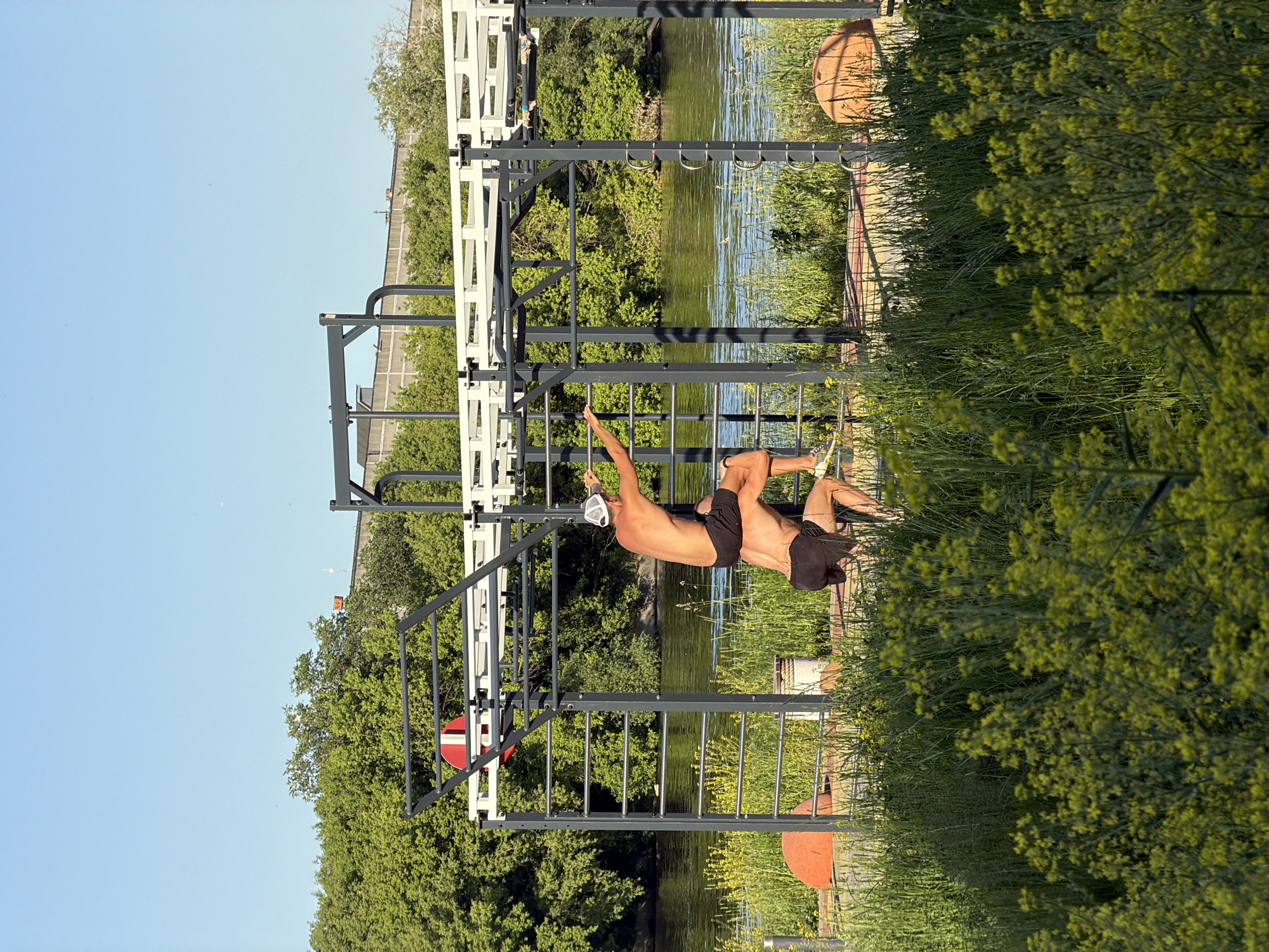 The image depicts an outdoor calisthenics park situated alongside a body of water, likely the Moskva River, within Moscow, Russia. The scene is bathed in bright sunlight under a clear blue sky.
In the foreground, lush green grass interspersed with numerous bright yellow wildflowers occupies the lower portion of the frame. Beyond this natural growth, a reddish-brown paved area, possibly a specialized rubberized surface, is visible, forming the base of a modern outdoor fitness rig. Two rounded, reddish-brown objects, possibly large stones or decorative elements, flank this paved area.
The central subject is a multi-sectioned fitness rig constructed from dark grey and white metal bars. A red and white circular target or sign is affixed to the upper left section of the rig. Two shirtless males, both wearing dark athletic shorts, are engaged in an acrobatic or advanced calisthenics maneuver on the rig. One male, wearing a white baseball cap, stands on the shoulders of the second male. The lower male is in a bent-knee stance, supporting the weight of the individual above him. The upper male is reaching upwards with his right hand, gripping one of the horizontal bars of the rig.
Behind the fitness park, a calm body of water extends horizontally across the mid-ground. On the opposite bank, a dense line of vibrant green deciduous trees forms a natural barrier. Above the tree line, a long, grey concrete structure, possibly a bridge, an elevated highway, or a railway line, is visible against the clear sky.