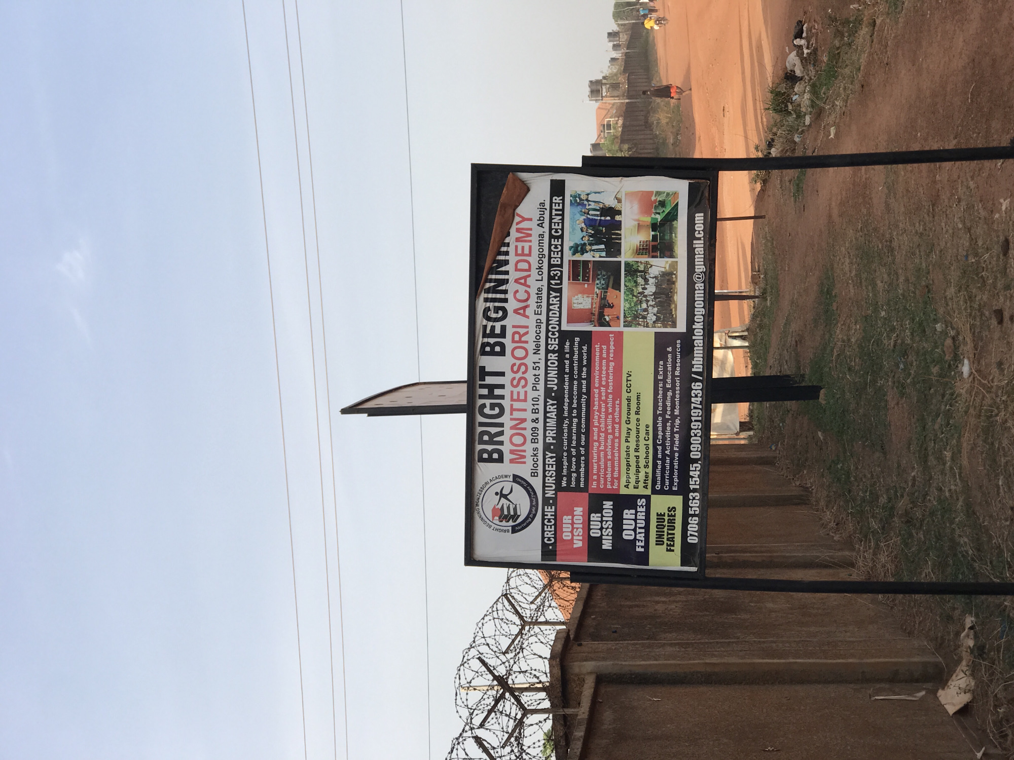 A large outdoor sign for "Bright Beginning Montessori Academy" is prominently displayed on the side of a dirt road in what appears to be a semi-urban area of Abuja, Nigeria. The sign, supported by metal poles, is the central focus of the image.
The sign details the school's name, "BRIGHT BEGINNING MONTESSORI ACADEMY," and its location: "Blocks B09 & B10, Plot 51, Nelocap Estate, Lokogoma, Abuja." It advertises programs including "CRECHE - NURSERY - PRIMARY - JUNIOR SECONDARY (1-3) BECE CENTER." The school's vision is to inspire "curiosity, independent and a life-long love of learning to become contributing members of our community and the world," while its mission is to foster self-esteem, problem-solving skills, and respect.
Highlighted features on the sign include an "Appropriate Play Ground," "CCTV," an "Equipped Resource Room," and "After School Care." Unique features listed are "Qualified and Capable Teachers," "Extra Curricular Activities," "Feeding," "Education & Explorative Field Trip," and "Montessori Resources." Contact phone numbers (0706 563 1545, 09039197436) and an email address (bbmalokogoma@gmail.com) are also visible. A school logo, depicting a stylized figure nurturing a plant or child, is present on the sign.
The setting includes a wide, reddish-brown dirt road in the background, where several people are seen walking in the distance. To the left of the sign, a concrete fence topped with coiled barbwire is visible. Overhead, power lines stretch across a clear, bright sky, indicating it is daytime, likely late morning or early afternoon. The overall scene reflects an outdoor, developing environment consistent with the location context of Abuja, Nigeria.