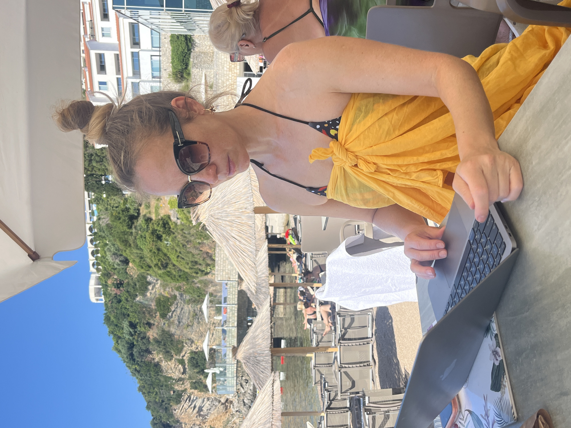 A woman with blonde hair pulled into a bun is seated at a table outdoors, working on a laptop. She is wearing large sunglasses and a yellow sarong tied around her chest over a bikini top. In the background, several thatched umbrellas and lounge chairs are visible on a beach, with people relaxing. The sea is calm and the sky is clear blue. Behind the beach, a hillside is covered in greenery and a white building with arched windows is situated on top. This scene is at Mogren Beach in Budva, Montenegro.