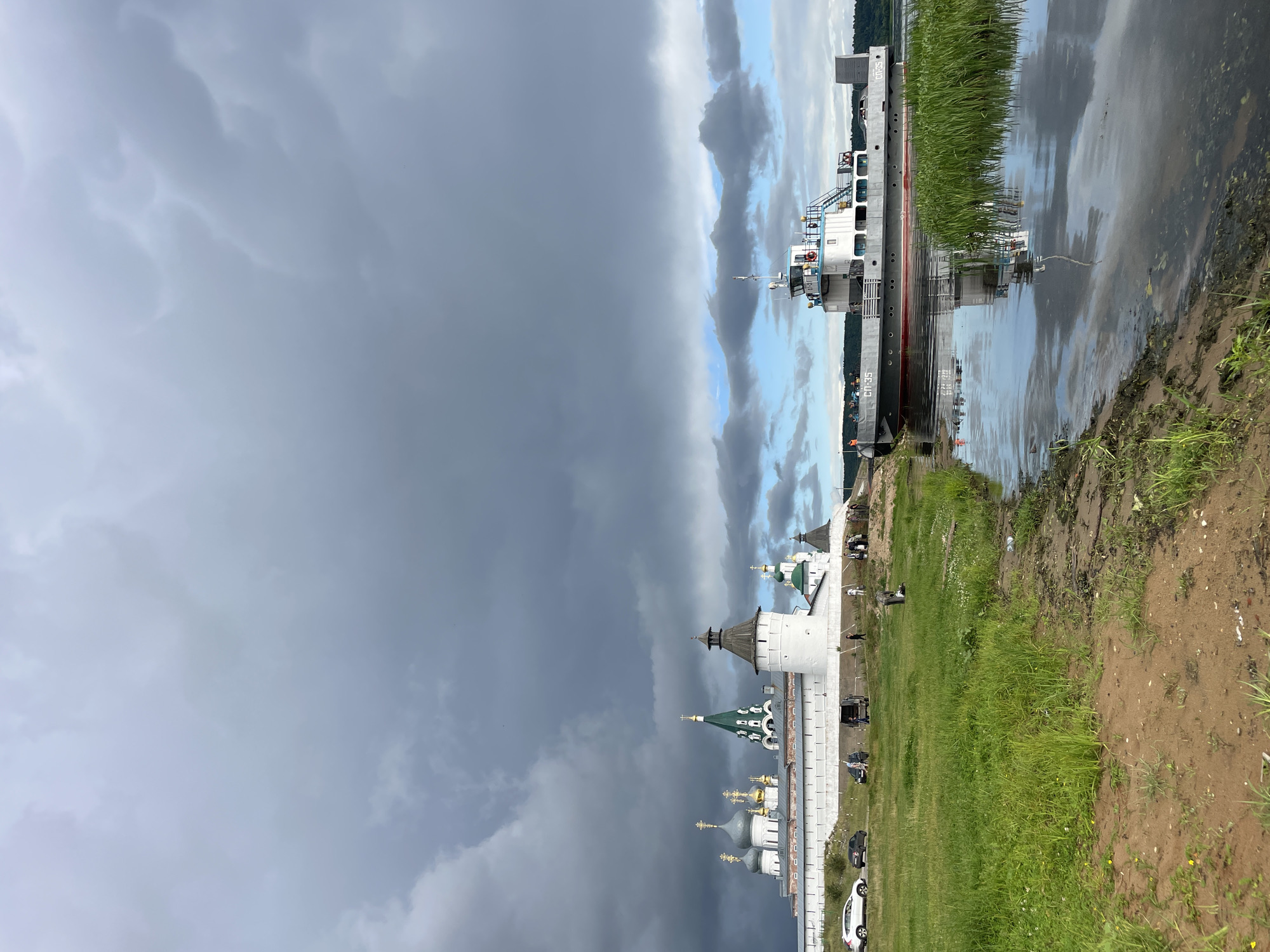 The image depicts a wide river with a ferryboat docked near the shore on the right side. On the left bank, a large, white fortress-like structure with multiple green and gold-domed buildings stands on a hill. Several cars are parked on a dirt road in front of the fortress. The sky is overcast with dark, dramatic clouds, though patches of blue sky are visible. The foreground shows a sandy riverbank with lush green grass growing along the water's edge. The reflection of the ferryboat and the sky is visible on the calm surface of the river. The location is identified as Makar'evo, Russia.
