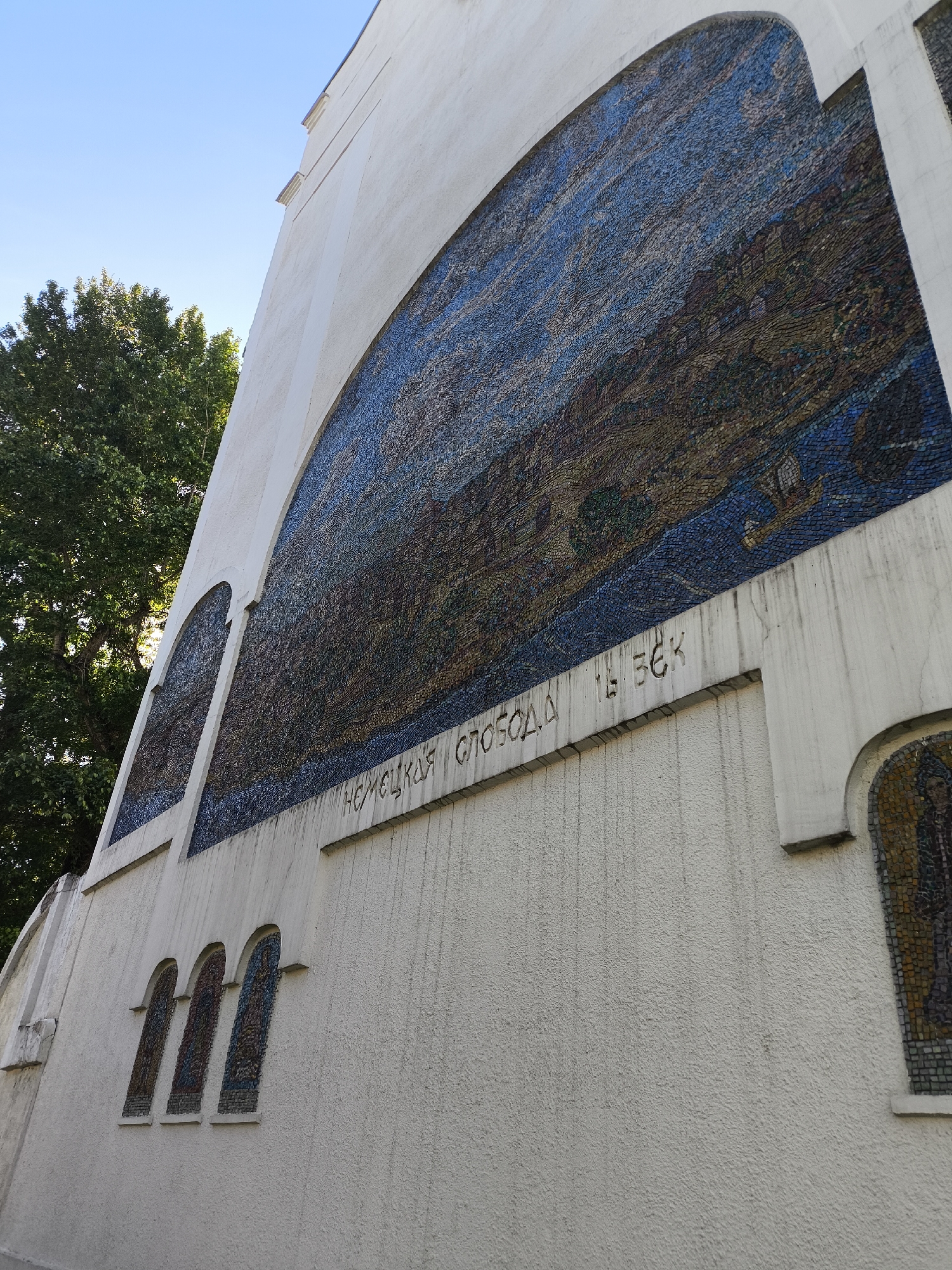A low-angle, medium shot captures the façade of a light-colored building featuring a mosaic and Russian text. The left side of the building is visible, with the mosaic forming a large arch at the top and several smaller arch-shaped mosaics below. The large mosaic displays a landscape in blue and brown hues, made up of numerous small tiles.
Beneath the large mosaic, a raised horizontal band bears an inscription in Russian: "НЕМЕЦКАЯ СЛОБОДА XVI ВЕК".
The building’s exterior is textured, with subtle vertical streaks of grime running down the surface. The environment includes a tree with dense green foliage, visible to the left of the building against a clear blue sky. The location is Unknown, Unknown.