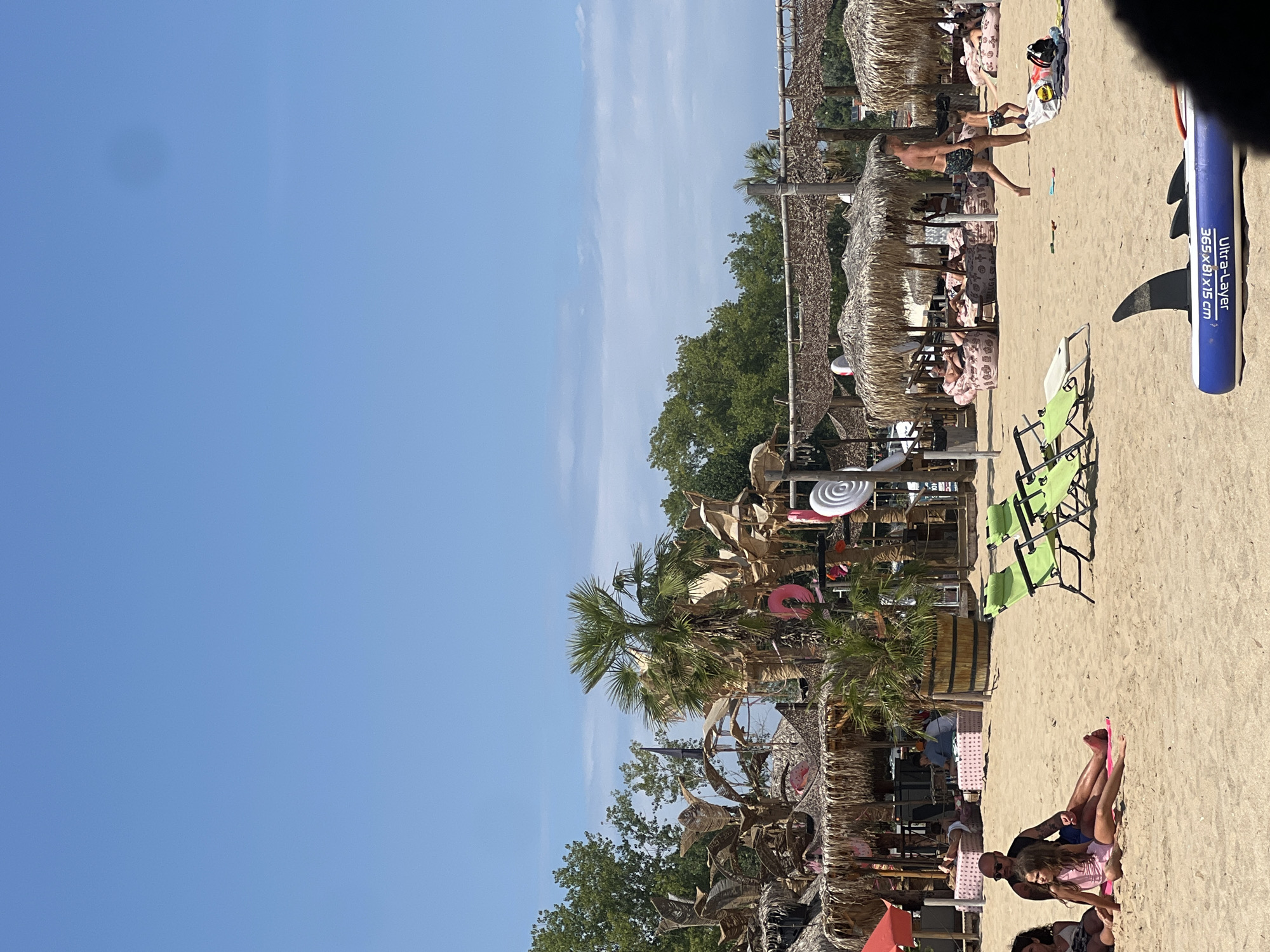 This is a photograph of a crowded beach with a clear blue sky. In the foreground, a family is relaxing on the sand, with a man and a young girl sitting close together, and another woman to their left. Two green lounge chairs are set up nearby. Further back, a man walks with a young child. The beach features numerous thatched-roof umbrellas providing shade, with people lounging on beanbag chairs underneath. The scene suggests a lively, resort-like atmosphere, typical of a summer day in Sozopol, Bulgaria.