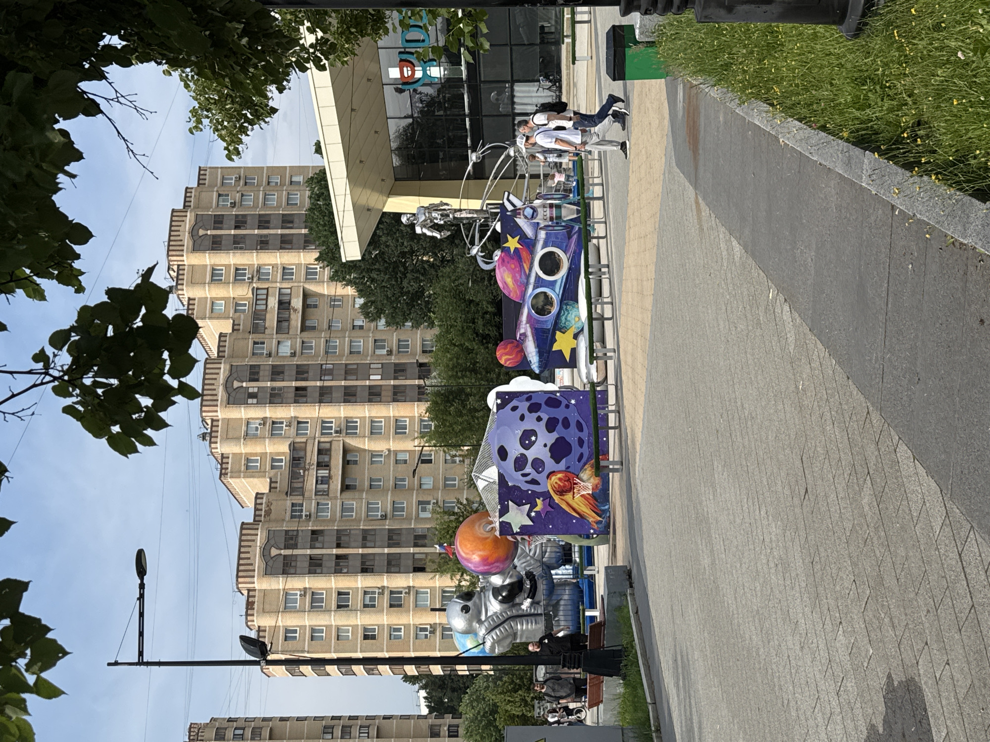 The image shows an outdoor scene in Zelenograd, Russia, featuring a public space with art installations and residential buildings in the background. In the foreground, there is a paved walkway with several large, colorful art pieces themed around space exploration. One installation depicts a rocket ship with planets and stars, another features a large astronaut figure, and a third shows a robotic sculpture on a platform. Several people are walking in the area. Behind the art installations, there are modern, multi-story apartment buildings with beige facades. Trees and greenery are visible throughout the scene, and there are streetlights and benches. A building with a glass facade and a visible logo is situated to the right. The sky is blue with some clouds.