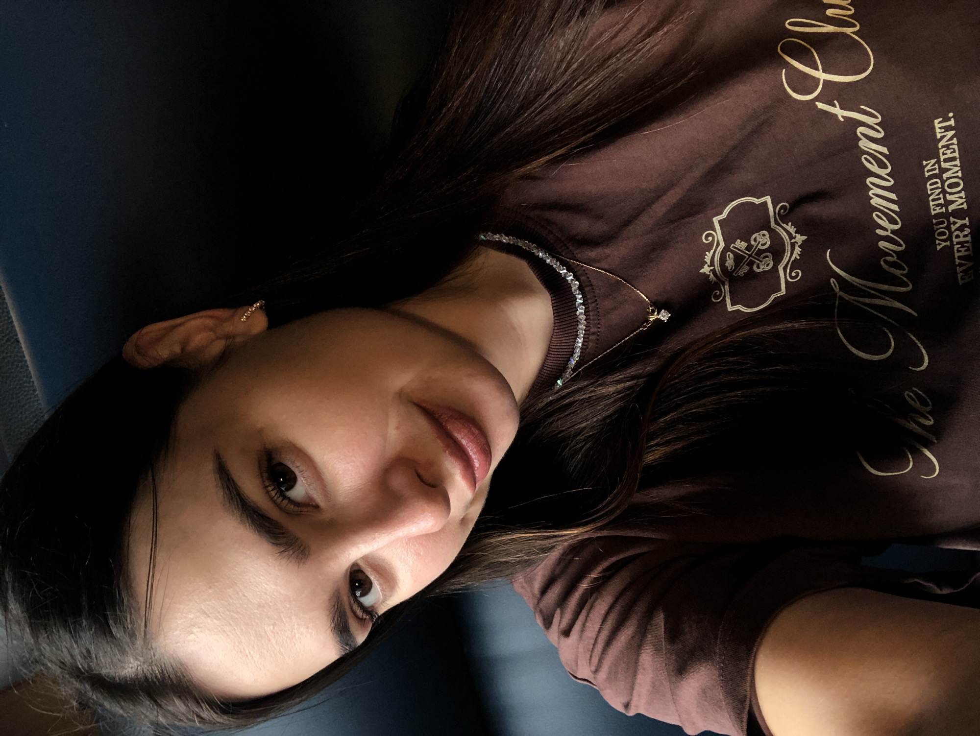 The image is a close-up selfie of a young woman with long, dark hair. She is wearing a brown t-shirt with a graphic design and text that reads "The Movement Clu YOU FIND IN EVERY MOMENT." She has a delicate necklace with a small pendant and a crystal choker. Her makeup includes lipstick and mascara. The background is dark and out of focus, suggesting an indoor setting. There is no information to suggest the location is in Izhevsk, Russia.