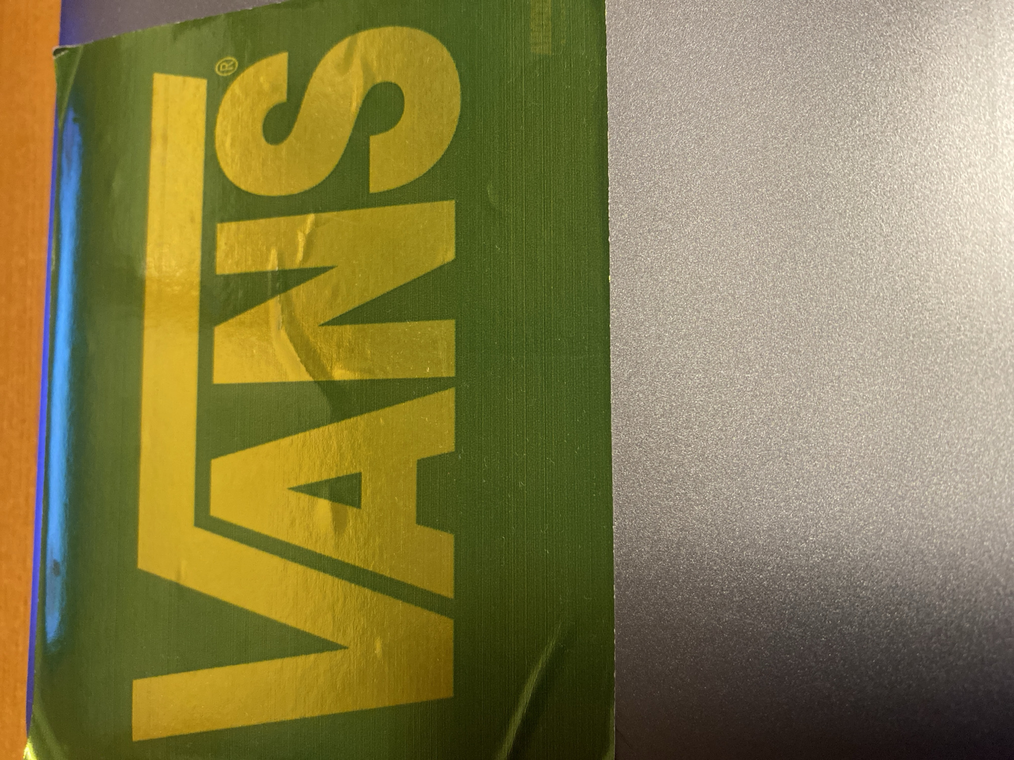 The image displays a rectangular green surface, likely a sticker or part of packaging, featuring the word "VANS" in a bold, yellow, sans-serif typeface. The letter "V" extends horizontally over the "ANS." A registered trademark symbol (®) is visible to the upper right of the "S." Both the green background and the yellow lettering exhibit a glossy, reflective quality, with notable highlights and some minor creasing or unevenness, particularly around the "V" and on the left side. The green surface has faint horizontal striations or texture visible.
The top edge of the green surface shows reflections of blue and purple light from an unseen source. Below the green surface, and extending across the bottom half of the image, is a flat, grey surface with a finely granular or speckled texture. This grey surface appears matte and slightly reflective, with a gradient from darker on the left to lighter on the right, suggesting a light source from the upper right. There are no people, explicit actions, or interactions visible in the frame. The environment is indistinct beyond the immediate surfaces shown.