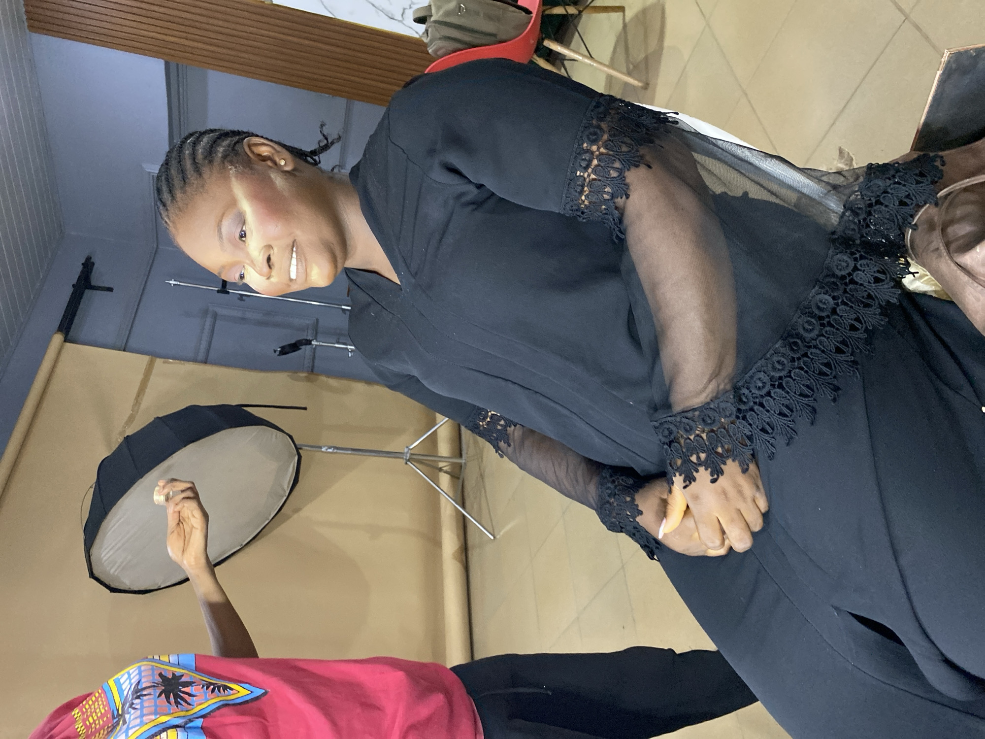 A woman with braided hair, wearing a black garment with lace sleeves, is seated indoors, presumably in a photography studio in Bukuru, Nigeria. The scene includes a softbox lighting setup positioned behind her. The woman is holding her hands together. A person wearing a red shirt stands close to the foreground. The environment seems to be set for a photoshoot.