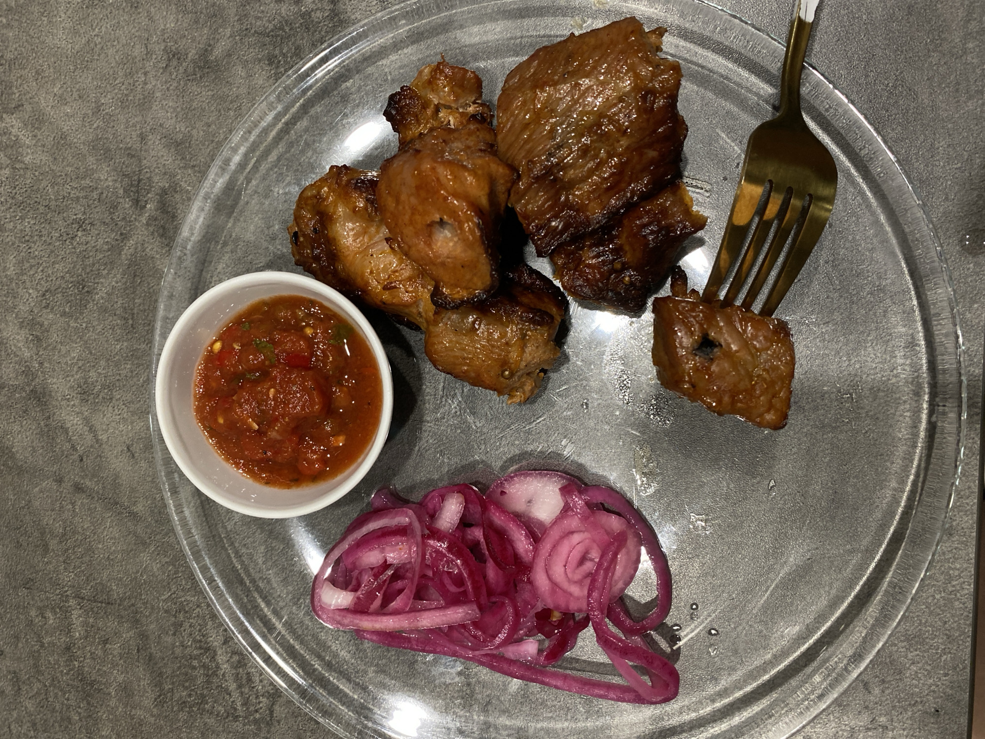 The image shows a plate of food, likely a meal, on a grey, textured surface. The plate appears to be made of clear glass or plastic. Food items include several pieces of cooked meat, possibly shashlik, a small white bowl of tomato-based sauce or relish, and a pile of marinated red onions. A golden fork is piercing one of the meat pieces. The scene appears to be set indoors, possibly in a restaurant or at a dining table. There is no visible indication that the image was taken in Sochi, Russia.