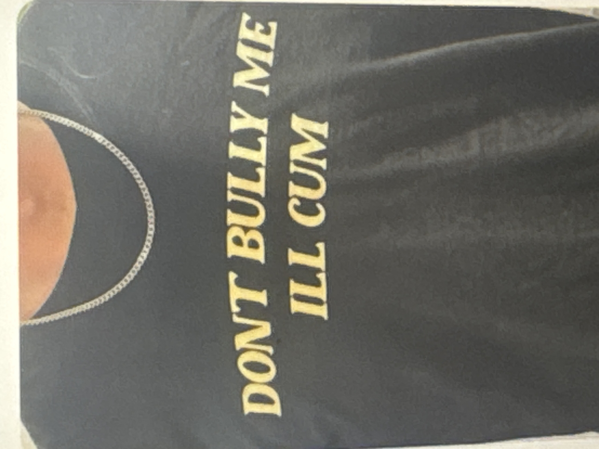 The image shows a dark t-shirt with yellow text printed vertically. The text reads "DON'T BULLY ME ILL CUM". A portion of a person's shoulder and neck, wearing a silver chain necklace, is visible on the left side of the image. The lighting appears to be artificial, and there are no clear indicators of the time of day, weather, or specific location beyond the provided context of Kuta Selatan, Indonesia. The image focuses on the t-shirt and its inscription.