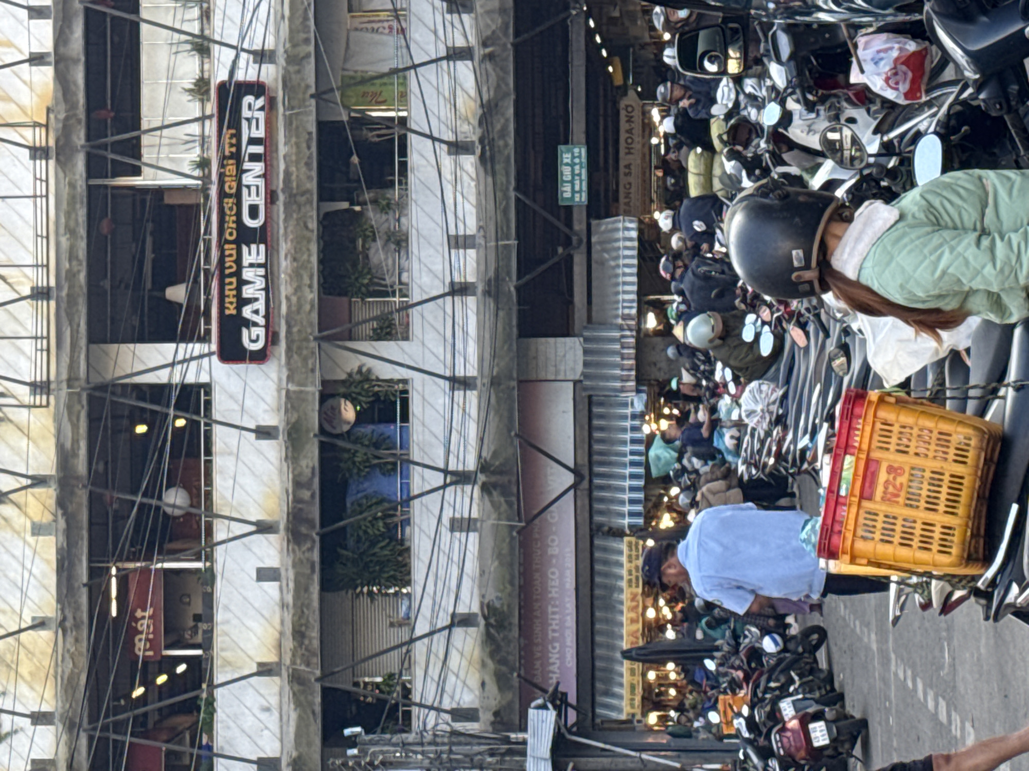 The image captures a street scene in Da Lat, Vietnam, likely near a market or commercial area given the presence of parked motorbikes and shops. In the foreground, a person wearing a light green quilted jacket and a dark helmet is partially visible, facing away from the camera. To their left, an orange plastic basket with red trim sits on a motorbike. Further back, a man in a light blue shirt and dark cap stands facing right, with many parked motorbikes behind him.
The background reveals a multi-story building with several balconies and shops. A prominent sign on the second floor reads "GAME CENTER" in white lettering on a black background, with Vietnamese text above it. Another sign, with pinkish-purple coloring, is visible on the first floor, displaying Vietnamese text related to meat products. Various shopfronts with awnings and bright lights are also visible, suggesting a bustling commercial environment. The scene is further cluttered with numerous overhead wires, common in Vietnamese urban settings. The overall impression is of a busy, everyday scene in a Vietnamese city.