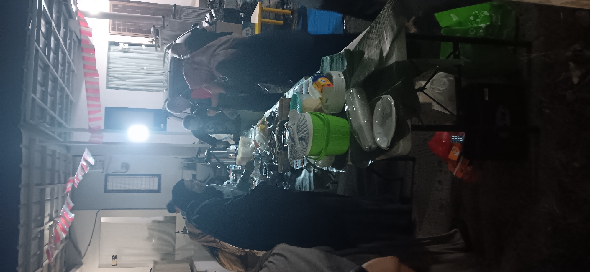 The image captures a nighttime outdoor gathering, likely a communal meal or celebration, in Depok, Indonesia. A long table is laden with numerous dishes of food, presented in various containers and arranged on platters. Several individuals, some wearing headscarves, are gathered around the table, with some appearing to be serving or selecting food. In the background, there's a car, a structure resembling a house with windows and curtains, and decorative red and white banners strung across an overhead structure. The scene is illuminated by artificial lights, including a bright spotlight. There are also green buckets, plates, and plastic-wrapped items on the table, suggesting preparation for or during a meal. Bags, possibly shopping or grocery bags, are visible on the ground near a folded table and chairs. The overall impression is one of a lively, informal social event.