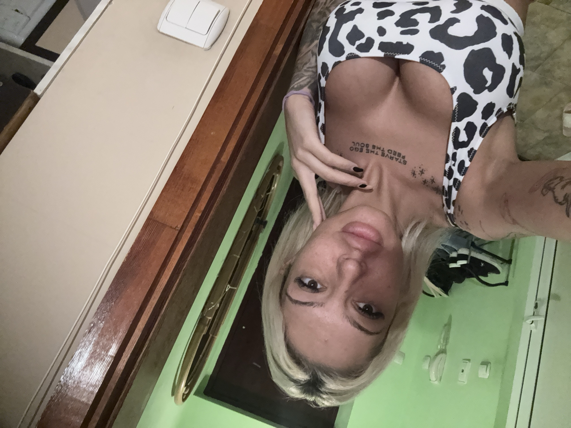 An indoor, vertically oriented selfie captures a woman with blonde hair, looking directly at the camera with a neutral expression. She is positioned towards the center-right of the frame. Her upper body is visible, wearing a form-fitting top with a black and white cow print pattern.
Notable details include tattoos on her upper chest, which read "STARVE THE EGO FEED THE SOUL" in capital letters, along with several small star-like symbols below the text. Another tattoo is partially visible on her right arm. Her left hand is raised towards her neck.
The background reveals interior elements: a light beige wall with a white light switch in the upper left, and a bright green wall to the right. A dark wooden trim or door frame runs vertically along the left side of the image, separating the beige and green walls. A gold-colored, circular decorative object hangs on the green wall, and another white light switch or outlet is visible on the green wall in the lower right. The overall setting suggests a casual indoor space in Plovdiv, Bulgaria.