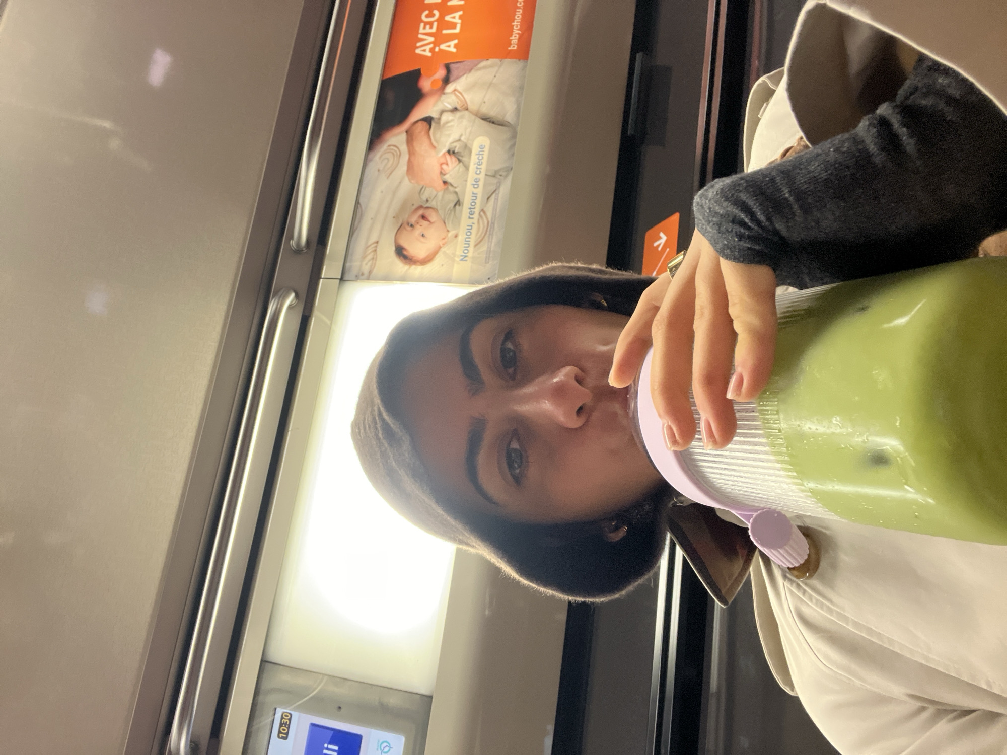 A woman is drinking from a large, pale green beverage container. She is wearing a brown hood and a light beige trench coat with a dark grey long-sleeved shirt underneath. The background appears to be the inside of a public transportation vehicle, possibly a train, with a light-colored wall featuring an advertisement for baby products in French. A digital display with a time of 10:30 is visible in the lower-left corner. The photograph is taken at an angle, with the subject holding the beverage up to her mouth.