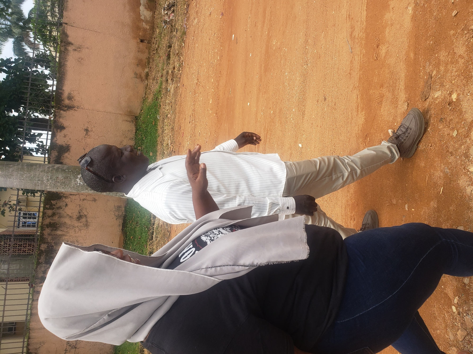 Two individuals are walking on a reddish-brown dirt path in Narayi, Nigeria. On the left, a person wearing a black t-shirt and dark blue jeans is covered from the head down with a light gray hijab that has a frayed edge. A graphic print is visible on the t-shirt. On the right, a man in a white striped long-sleeved shirt and light beige pants walks with sunglasses perched on his head. His left arm is extended, palm open, as if speaking or gesturing. Both are walking from left to right. The environment features a textured, light brown wall behind them, with some green vegetation and trees visible above the wall, as well as a tall, rough-barked tree trunk on the left. The ground is a loose dirt surface with scattered small stones and dried leaves.