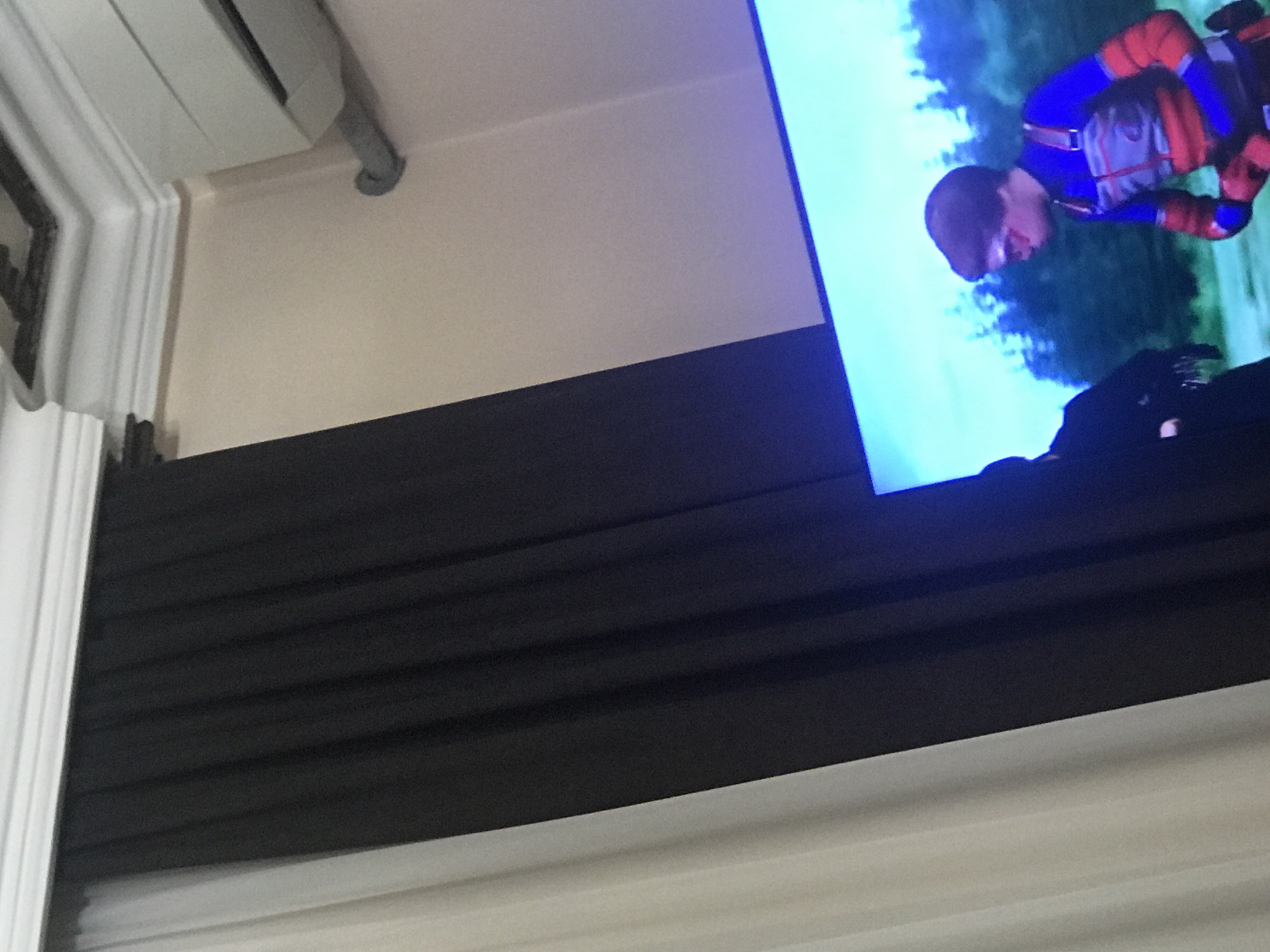The image shows a television screen displaying a person in a blue and red suit, likely an action or superhero costume, with a green, blurred background. The TV is mounted on a wall above what appears to be a dark-colored headboard or decorative wall paneling. Above the TV, there's a white ceiling with some sort of ventilation or AC unit and associated piping. The lower portion of the frame contains white curtains or drapery. The image doesn't provide enough information to pinpoint the specific location in Yerevan, Armenia.
