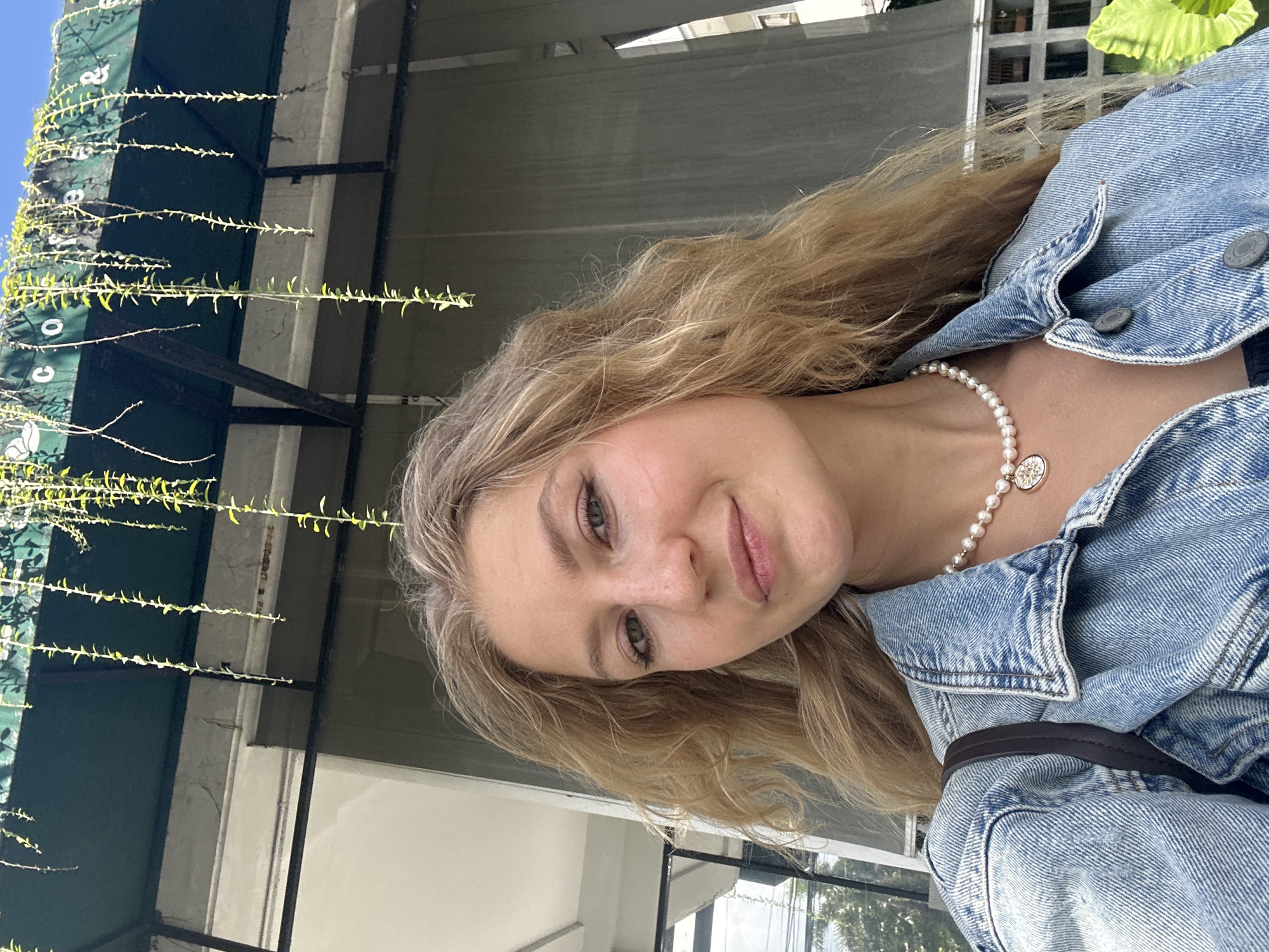 A woman with blonde hair, wearing a denim jacket and a pearl necklace with a gold pendant, is facing the camera. The background includes a building with a green awning, foliage, and structures suggestive of an outdoor setting. Given the prompt specifying the location as Kuta Selatan, Indonesia, the scene is likely an outdoor café or restaurant setting within the city.