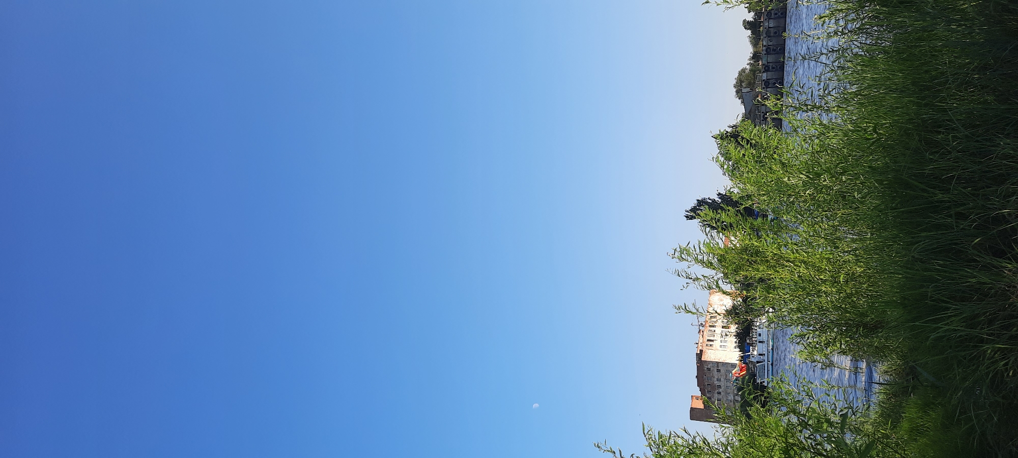 The image captures a serene daytime scene of a waterfront with lush green foliage in the foreground. In the mid-ground, a calm body of water reflects the clear blue sky. Across the water, the city of Astrakhan, Russia, is visible. A partially visible building, possibly an old or under-renovation structure, stands out with its brickwork and numerous windows. A white boat with a red flag is docked or moving near the shore. In the distance, beyond the building and boat, a structure resembling a dam or a series of piers is partially obscured by trees. A sliver of the moon is discernible in the vast, unblemished sky. The overall impression is one of a tranquil day by the river, showcasing a blend of natural beauty and urban infrastructure.