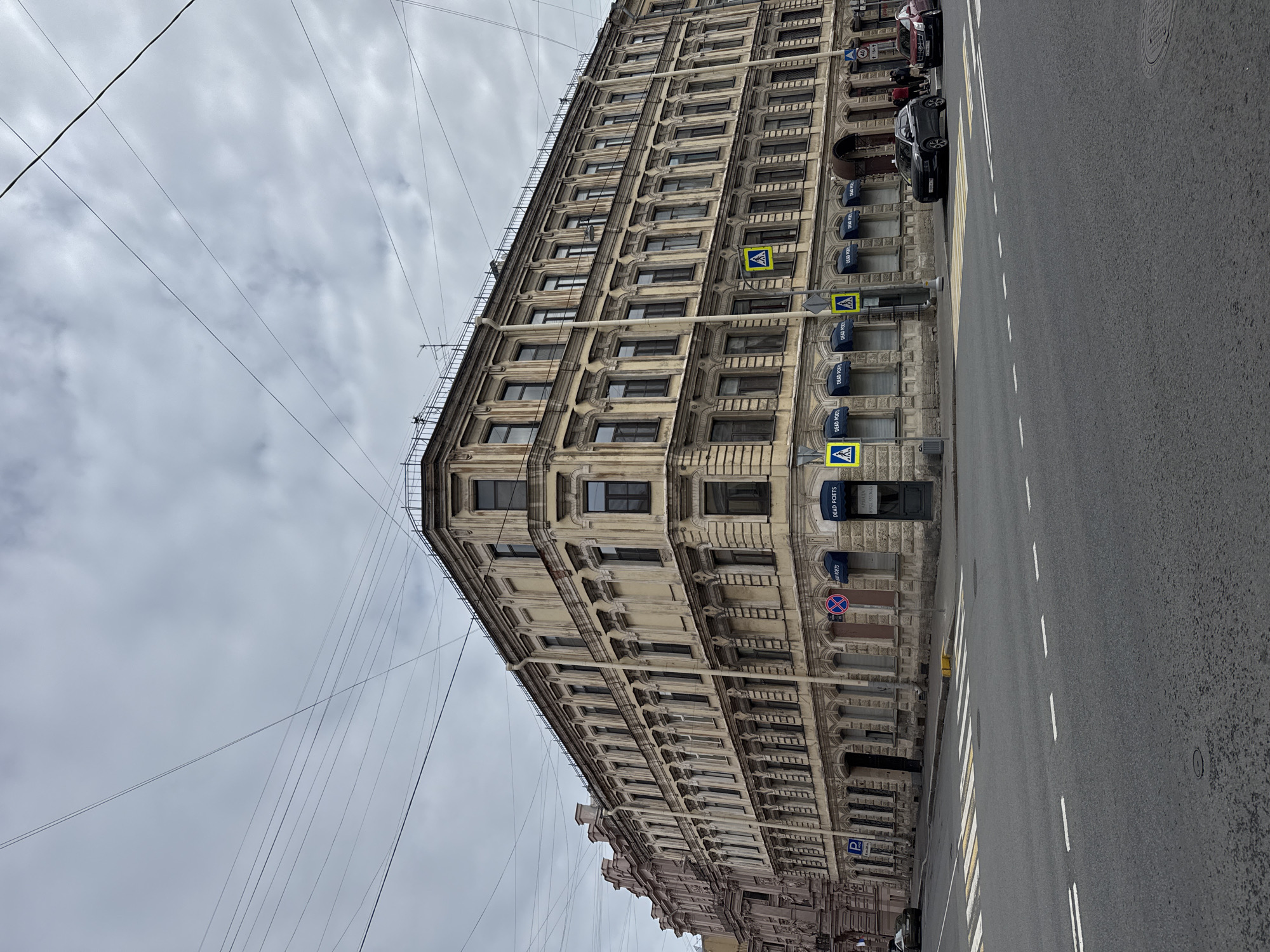 The image shows a tall, light-colored building in Saint Petersburg, Russia. The building has many windows and ornate architectural details. There are pedestrian crossing signs attached to the building's facade. A street runs along the side of the building, with cars parked along the curb. The sky is overcast. Power lines are visible in the sky.