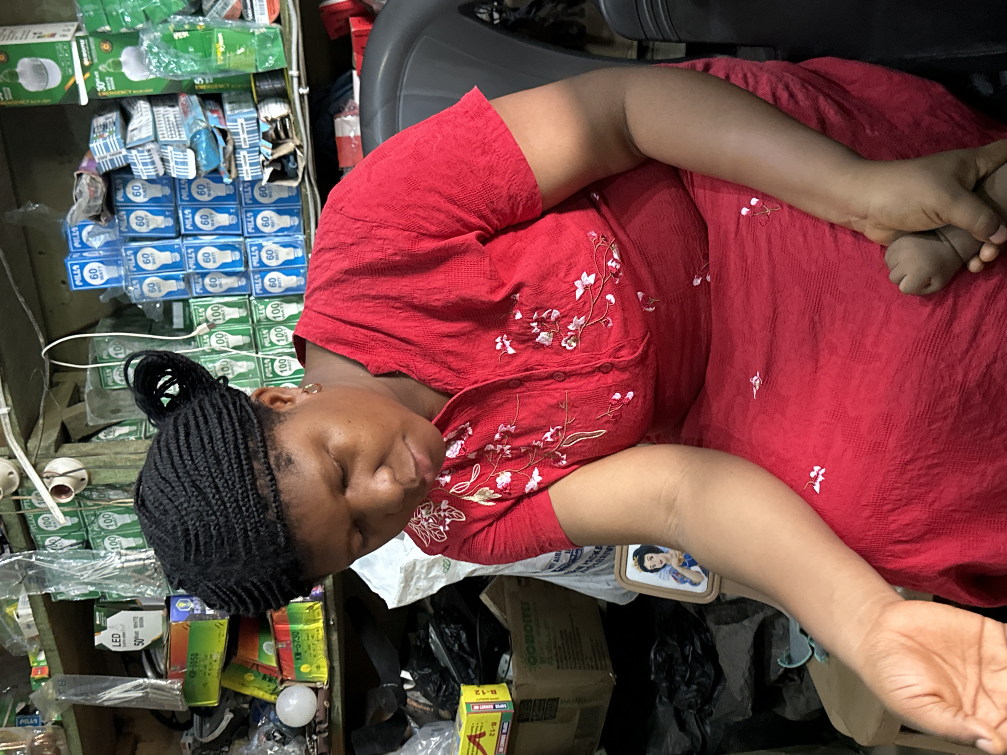 A Black woman wearing a red, embroidered dress is seated, looking down and to her left. Her braided hair is pulled up and back in a bun. The setting appears to be a shop in Narayi, Nigeria, with shelves stocked with various items, including boxes of light bulbs and what seem to be electrical components. A framed picture of Snow White is visible near her left arm. Her left arm is extended, and her hand is holding the tiny feet of an infant. The lighting is moderate, and the background is cluttered with merchandise and plastic bags.