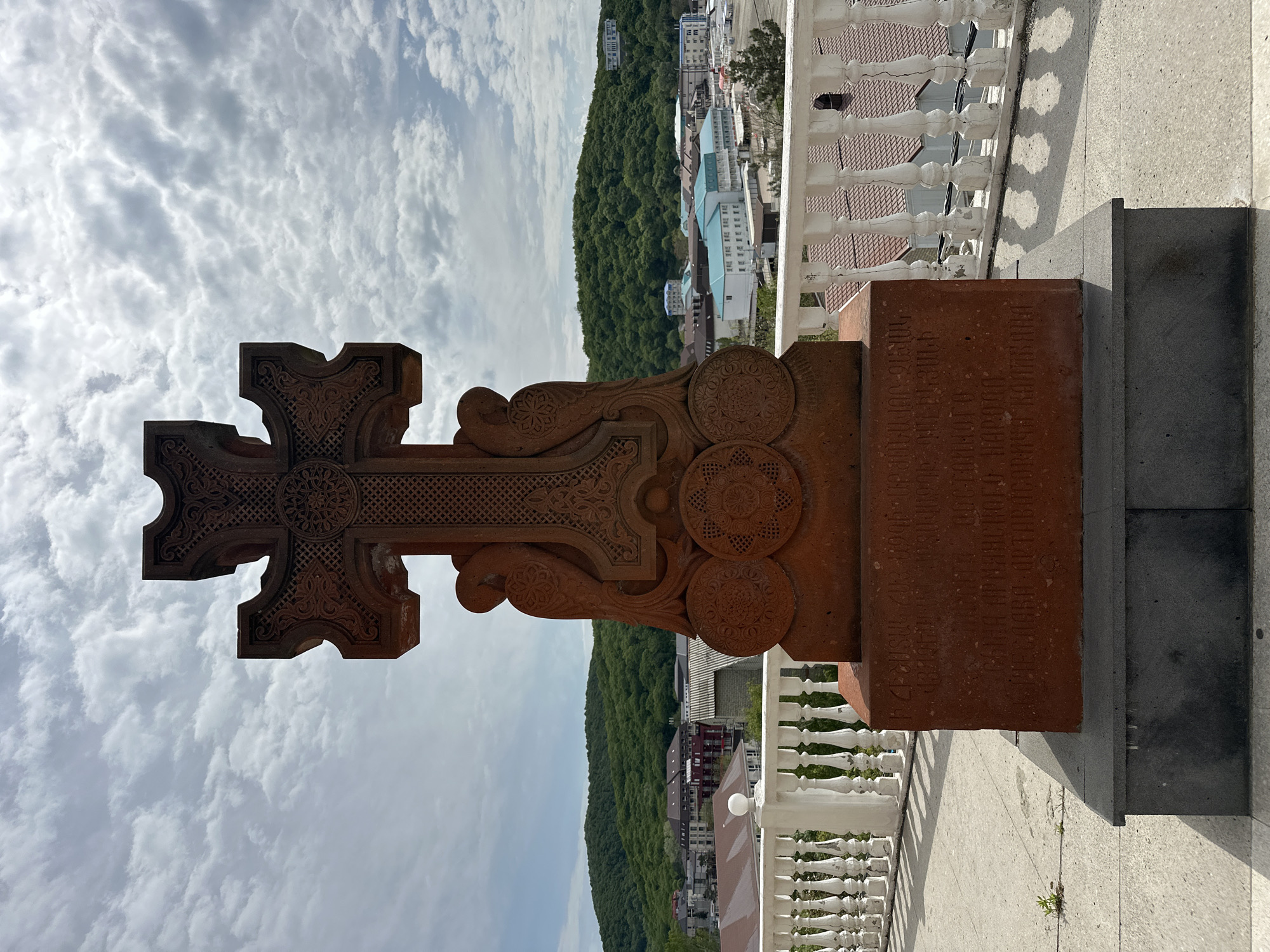 The image shows a vertical Armenian cross-stone (khachkar) in an outdoor setting. The cross is elaborately carved from reddish-brown stone and features circular patterns on the arms and base. The base has an inscription in Armenian script. Behind the cross-stone, a cityscape with a hillside covered in trees is visible. A white balustrade runs along the right side of the image, suggesting the cross-stone is located on a platform or terrace. The sky is partly cloudy. It is possible that the image was taken in the city of Novomikhaylovsky, Russia, however, this detail is not ascertainable by this image only.