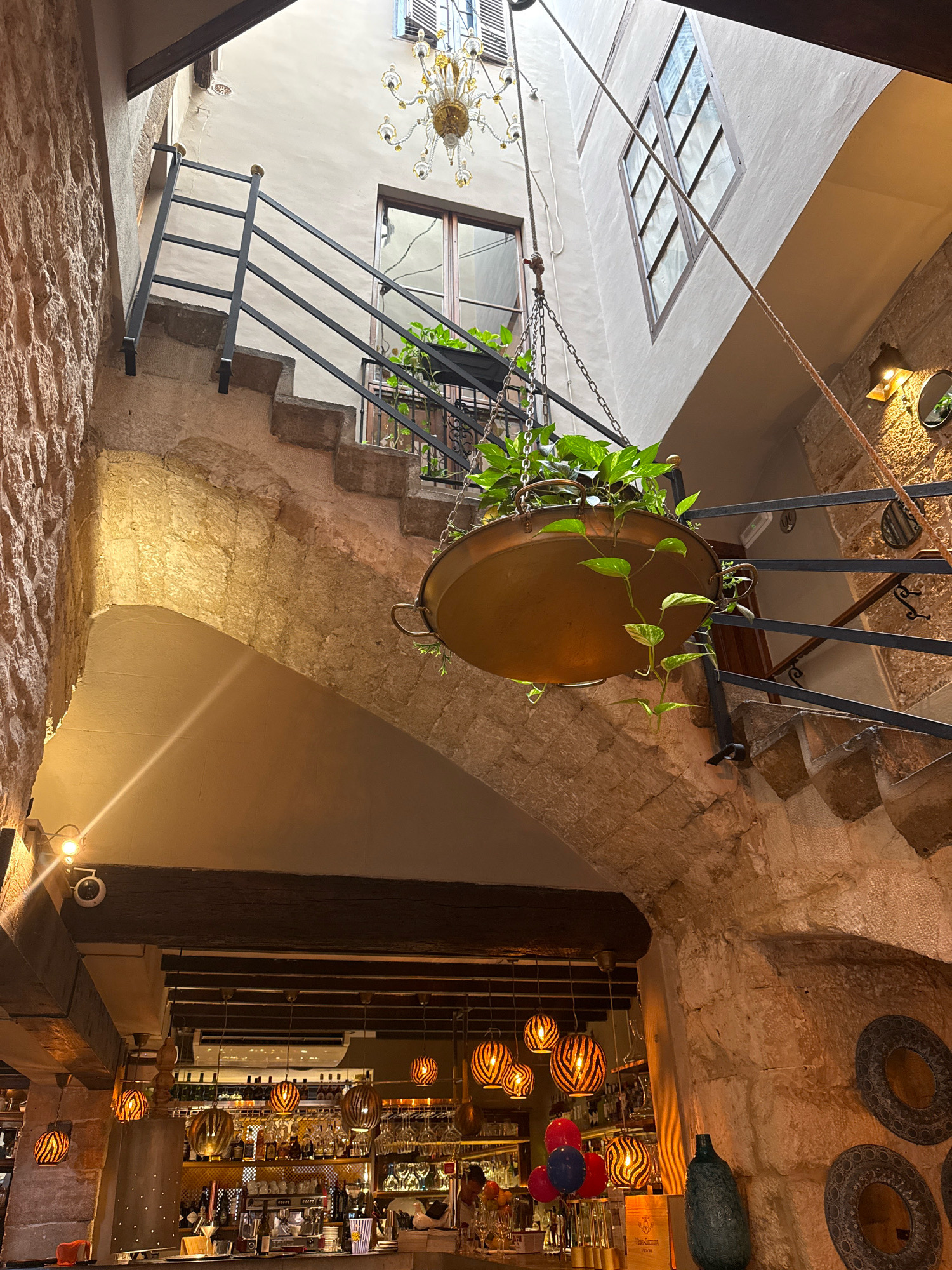 An interior shot captures a multi-level stone building with exposed stone walls and arched walkways. A metal staircase ascends on the left, leading to an upper level with a window and a decorative chandelier hanging from the ceiling. A large, copper hanging planter filled with green plants is suspended from chains in the center of the frame, casting a warm glow. On the right, another metal staircase railing is visible. Below the arches, a bar area is visible with shelves stocked with bottles and glasses, illuminated by striped pendant lights. A security camera is mounted on the ceiling above the bar. The overall atmosphere suggests a rustic yet elegant restaurant or bar interior. The location is identified as Palma, Spain.