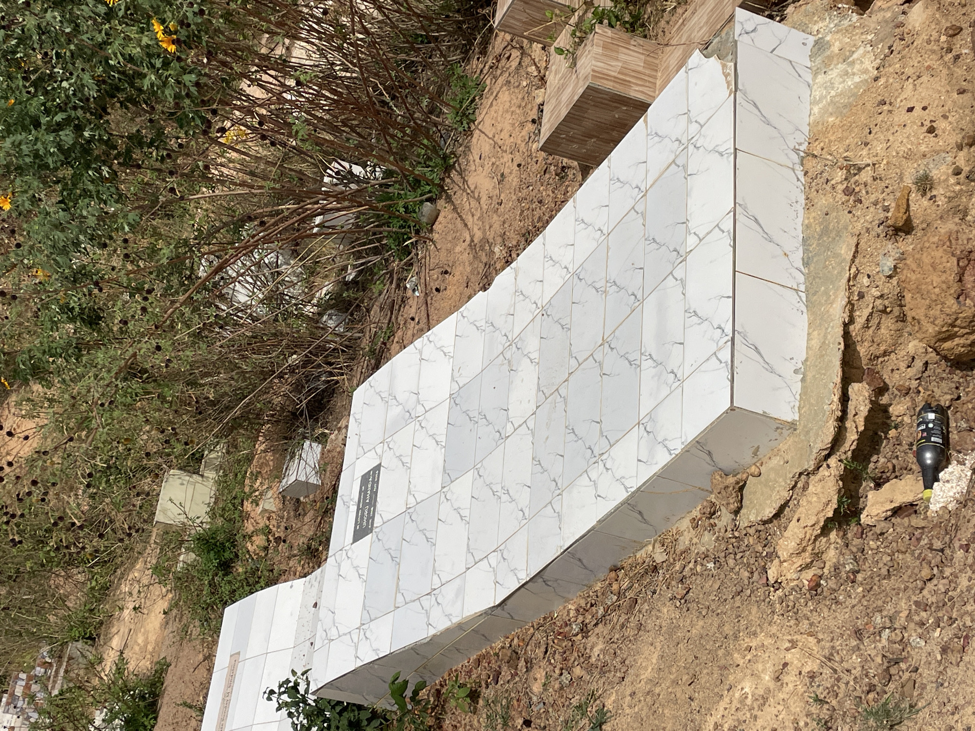 The image depicts a cemetery in Bar Jirgi Summa, Nigeria. A large, white marble-tiled tombstone dominates the scene. The tombstone appears newly constructed and features a small, dark plaque near its left edge. The surrounding ground is dry and dusty, interspersed with sparse vegetation and small rocks. A black plastic bottle is visible near the tombstone on the right. Other smaller tombstones are partially visible in the background.