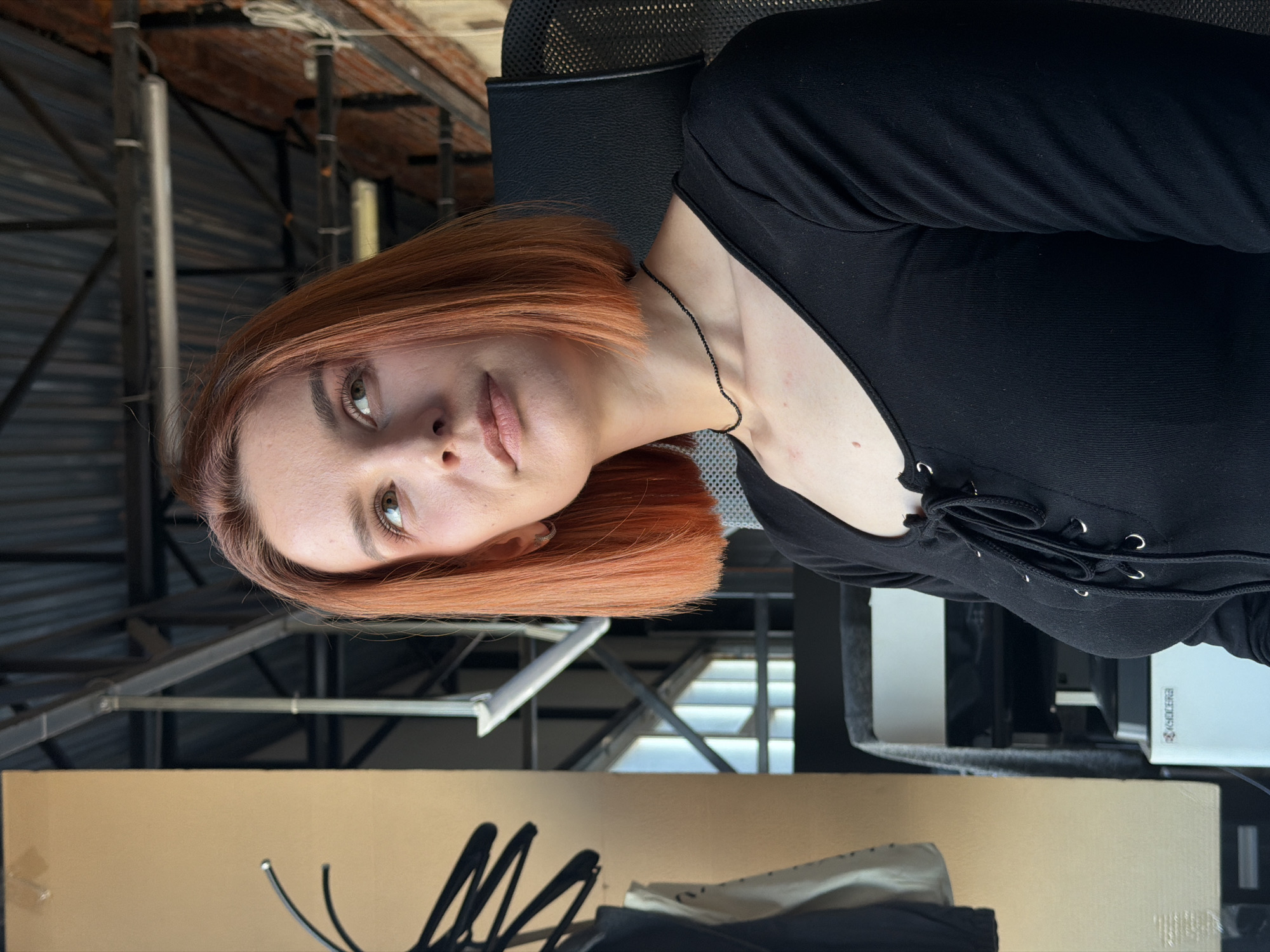 A rotated medium shot depicts a person with short, reddish-brown hair and fair skin. The person is wearing a black, long-sleeved top with lace-up detailing at the neckline. The person is seated in a black office chair. The background includes metal scaffolding, corrugated roofing, and a beige cardboard box. A white printer is positioned behind the person. Based on additional details, the location is consistent with Chaikovsky, Russia.