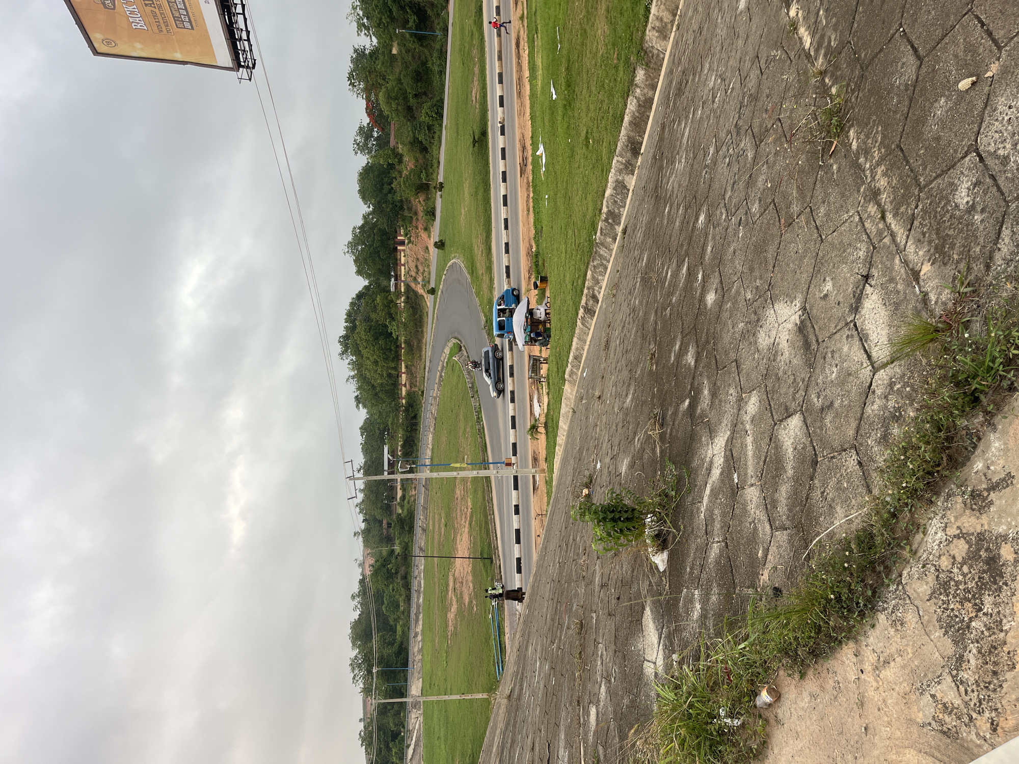 A tilted, wide-angle shot depicts a road curving through a green landscape in Osogbo, Nigeria. The road is paved and lined with alternating black and white markers. Several vehicles are visible: a blue car and a silver car. A small vendor stand with a white awning is adjacent to the blue car. A pedestrian in a red shirt walks along the road. The sky is overcast. A billboard is visible at the top left of the frame. The foreground shows a paved barrier with weeds and grass growing out of the cracks. Utility poles with power lines run parallel to the road. Trees are clustered around the perimeter of the grassy areas.